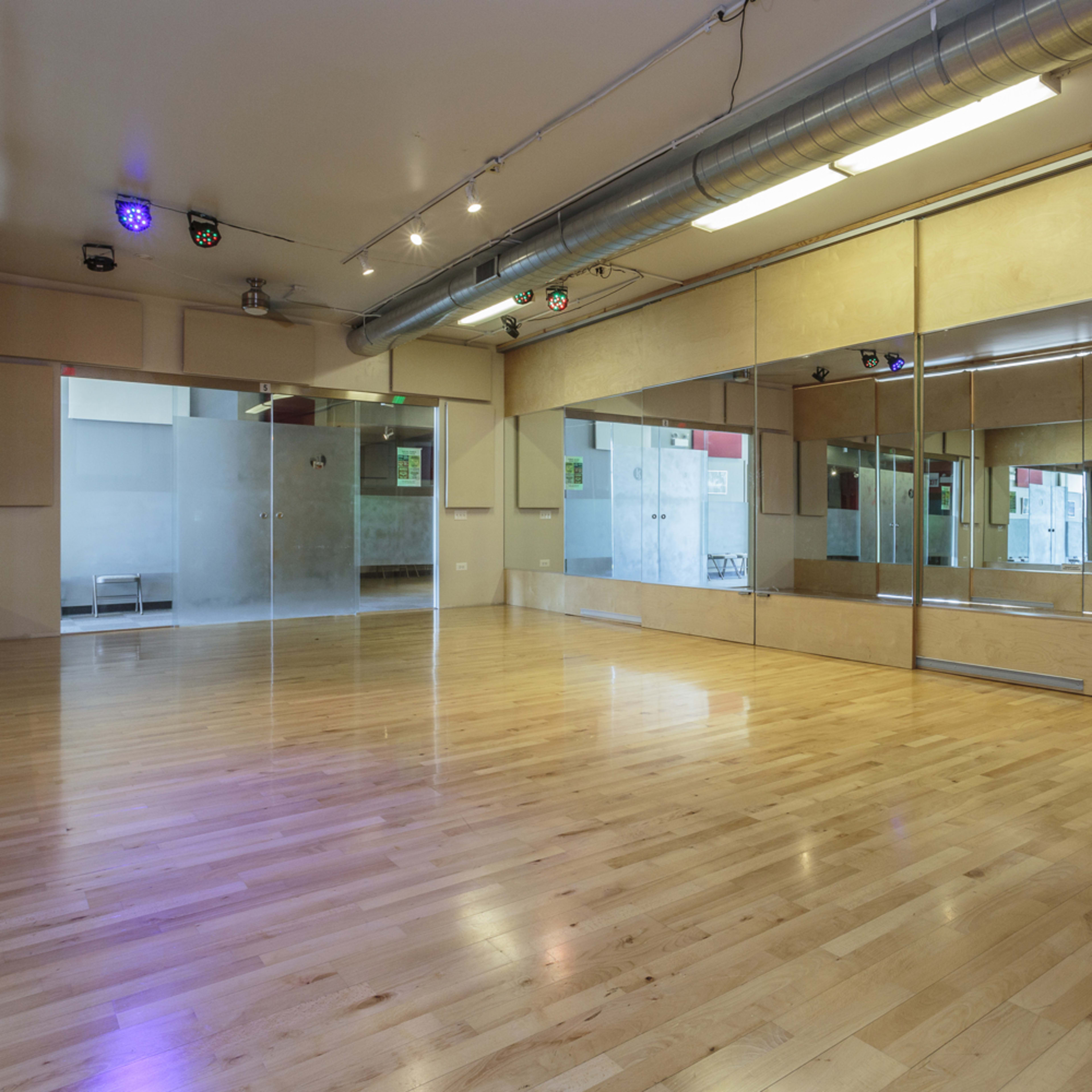A spacious dance studio with wooden flooring, mirrored walls, and overhead lighting.