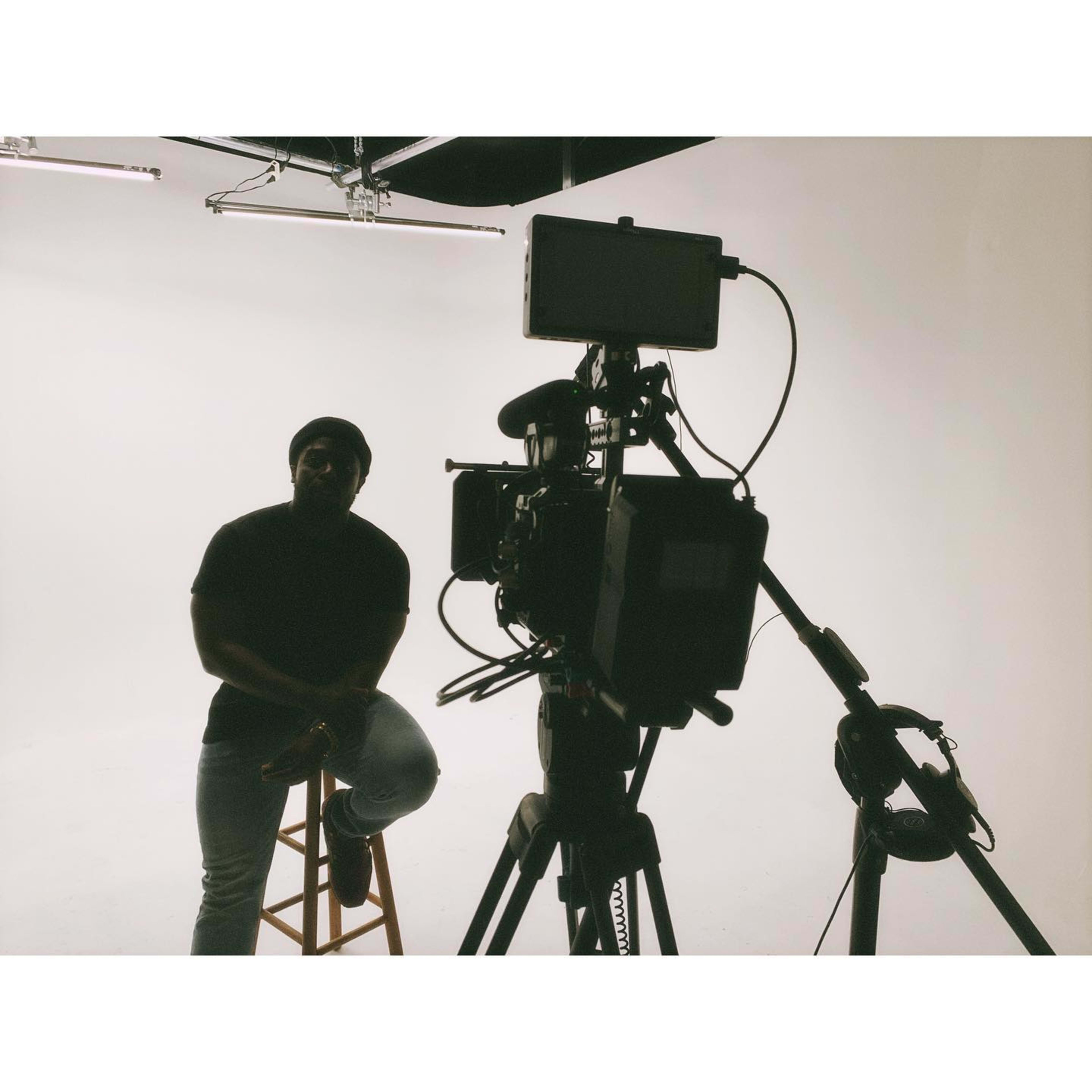 A person sits on a stool in front of a camera set up in a plain studio with bright lighting.