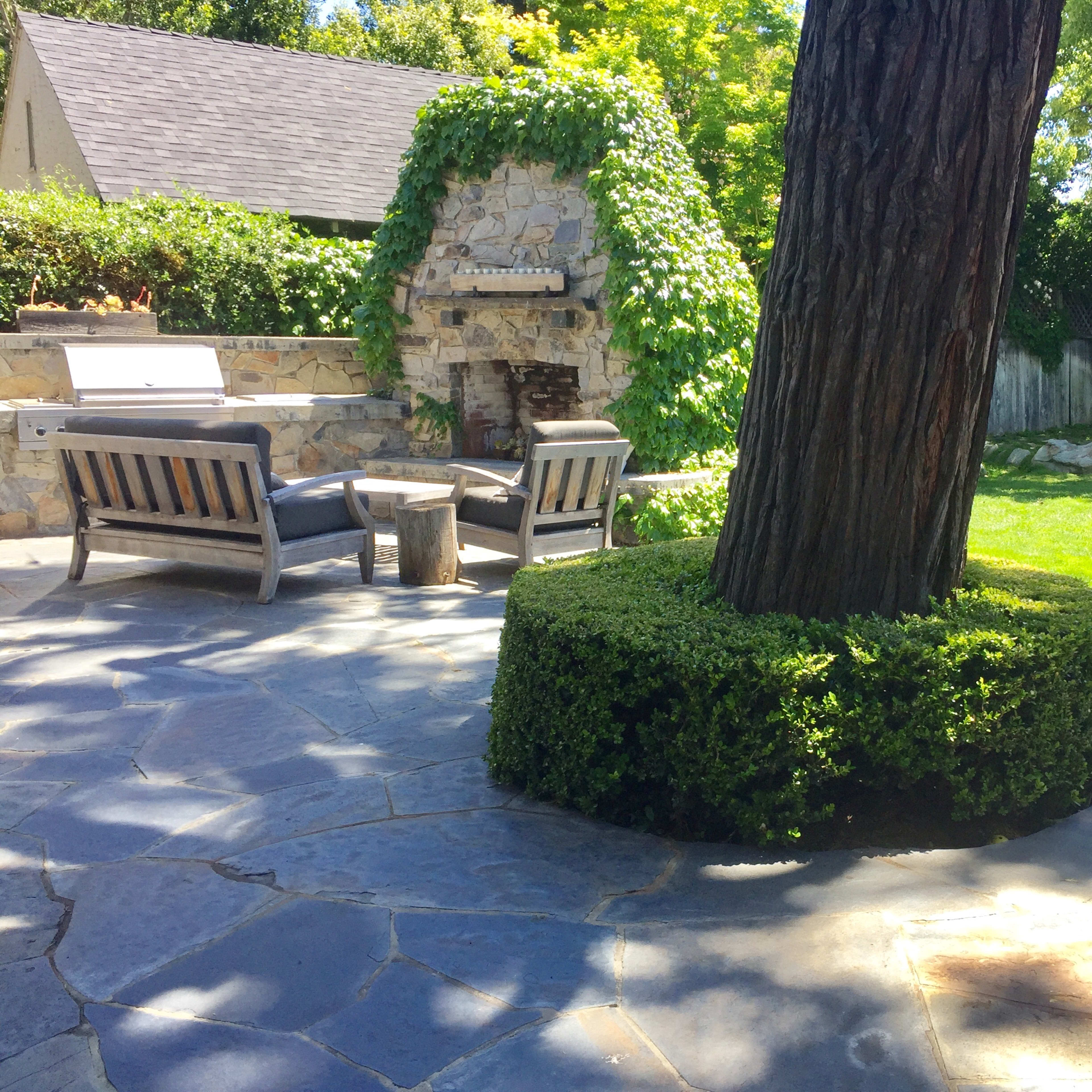 A stone patio features a large tree surrounded by a circular hedge, with seating and a stone fireplace in the background.