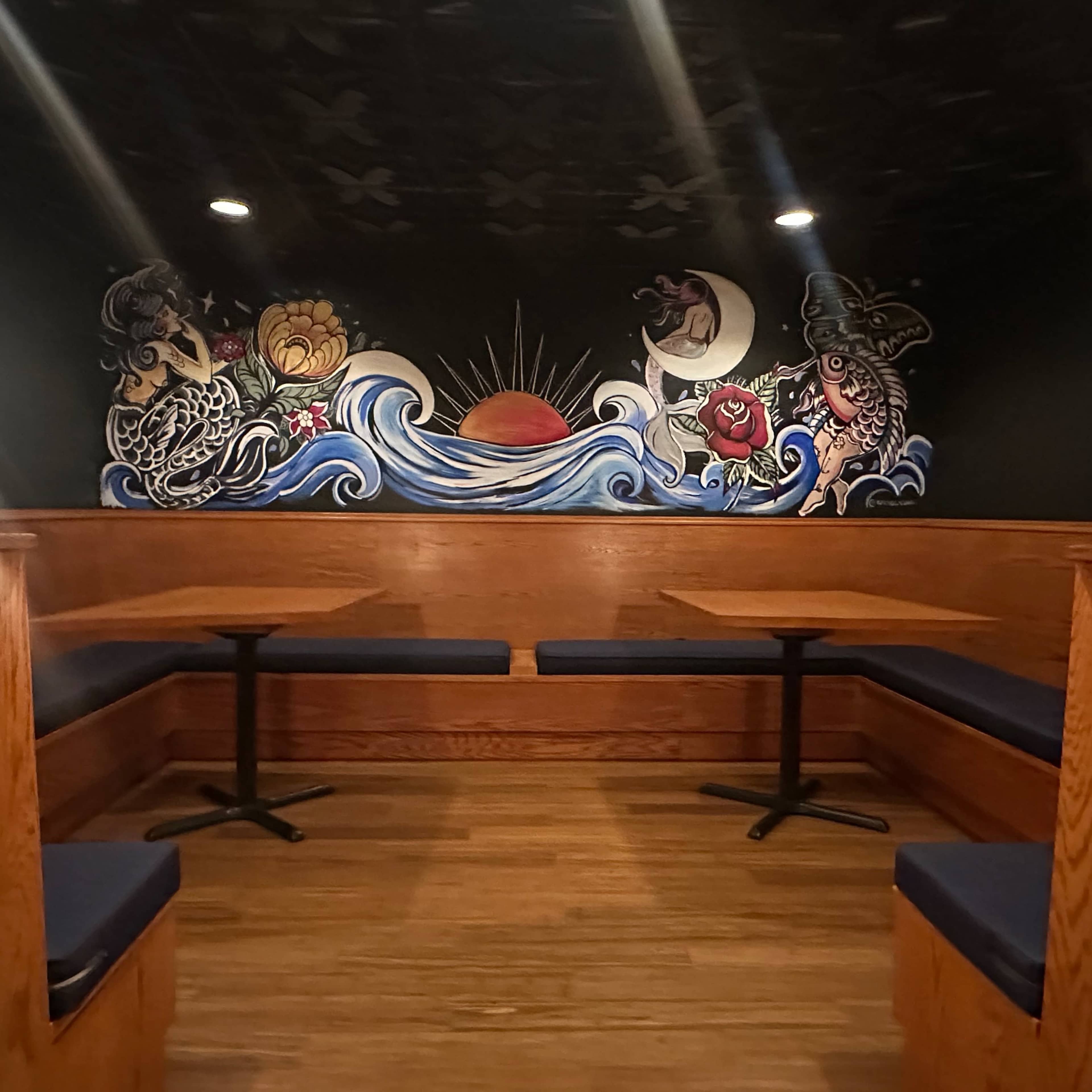 A cozy seating area in a dimly lit room featuring wooden booths and a colorful mural of waves and celestial elements on the wall.