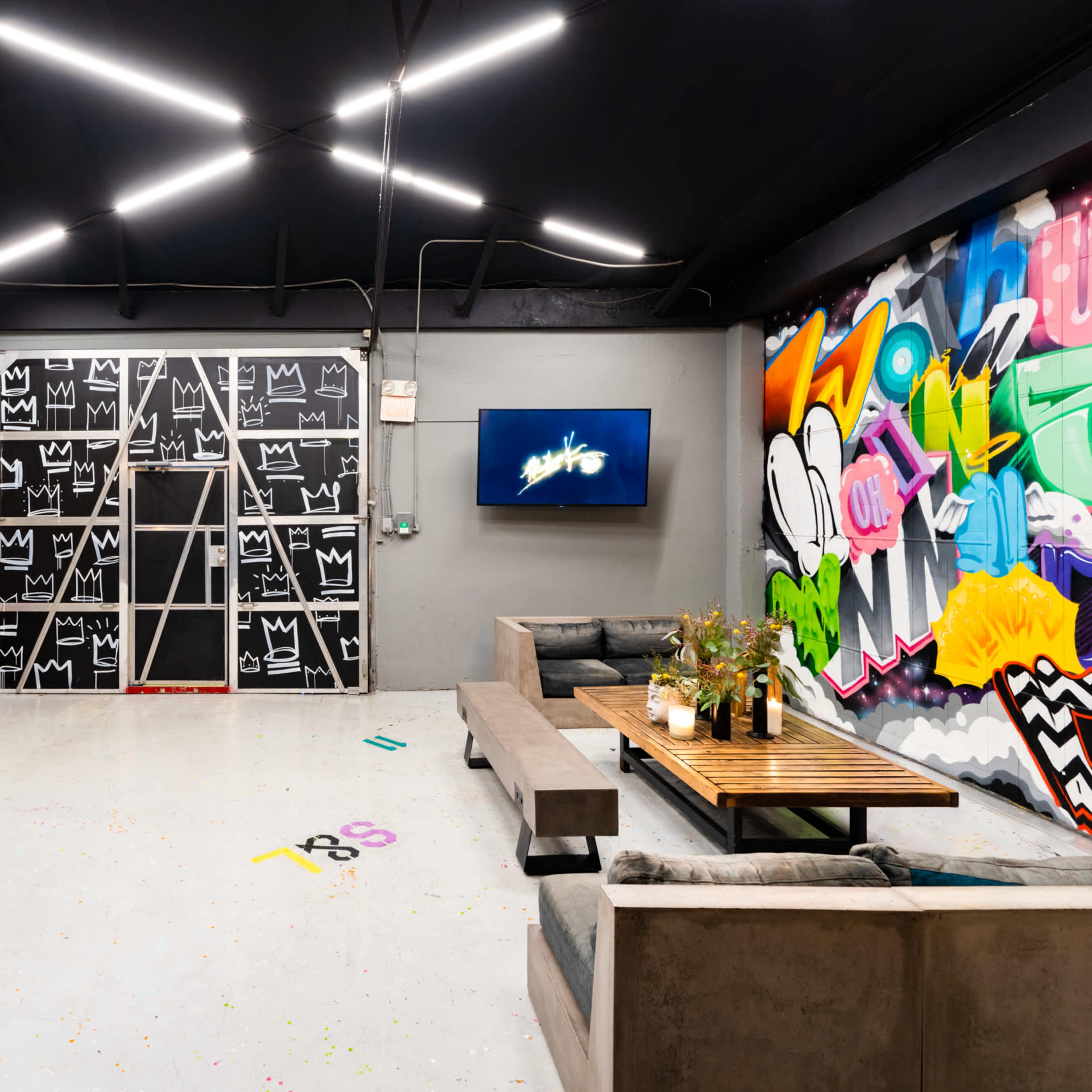 An industrial-style room featuring a colorful mural, a long concrete table with seating, a television on the wall, and a large blackboard-style door.