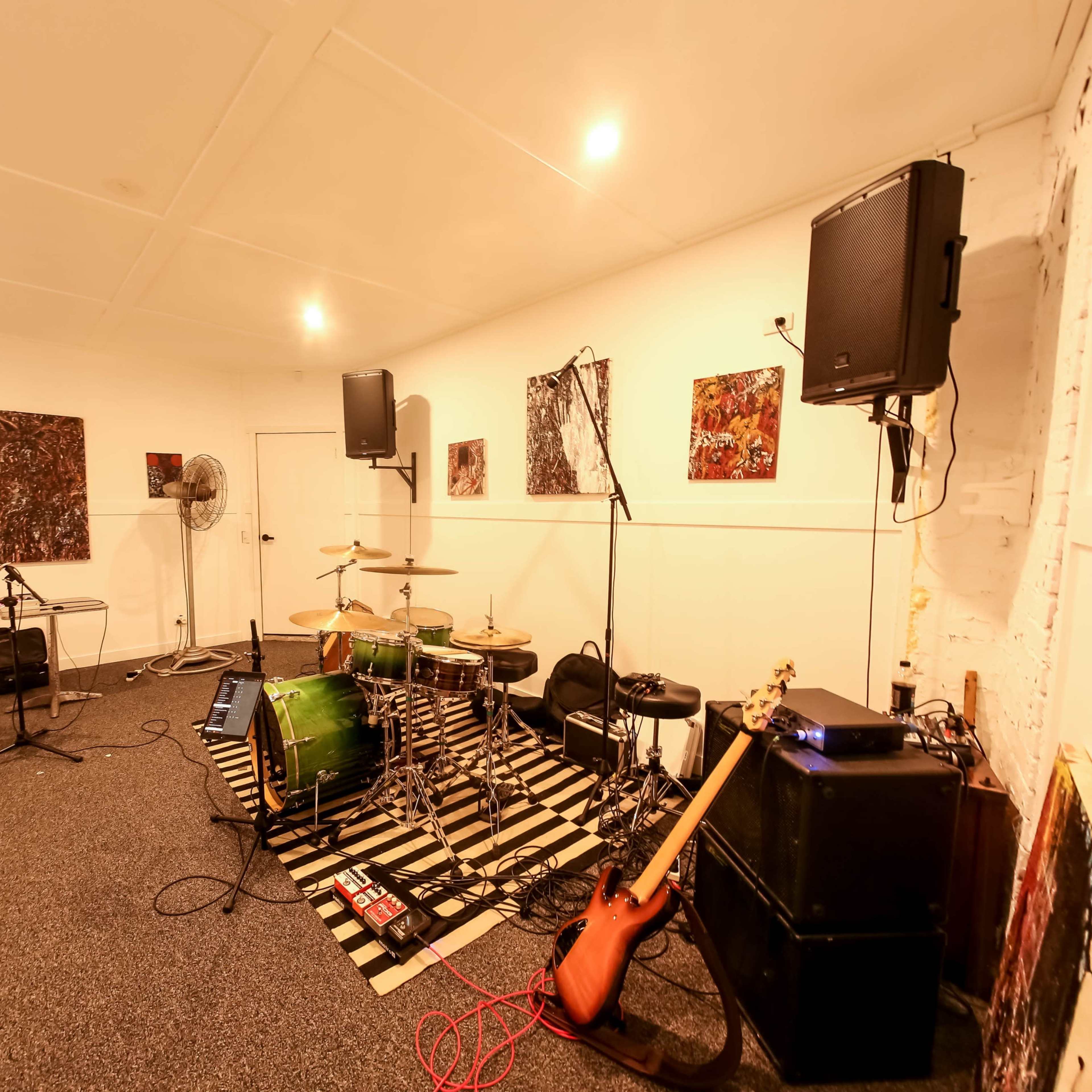 A music rehearsal space with a drum set, electric guitar, amplifiers, and sound equipment arranged along the walls.