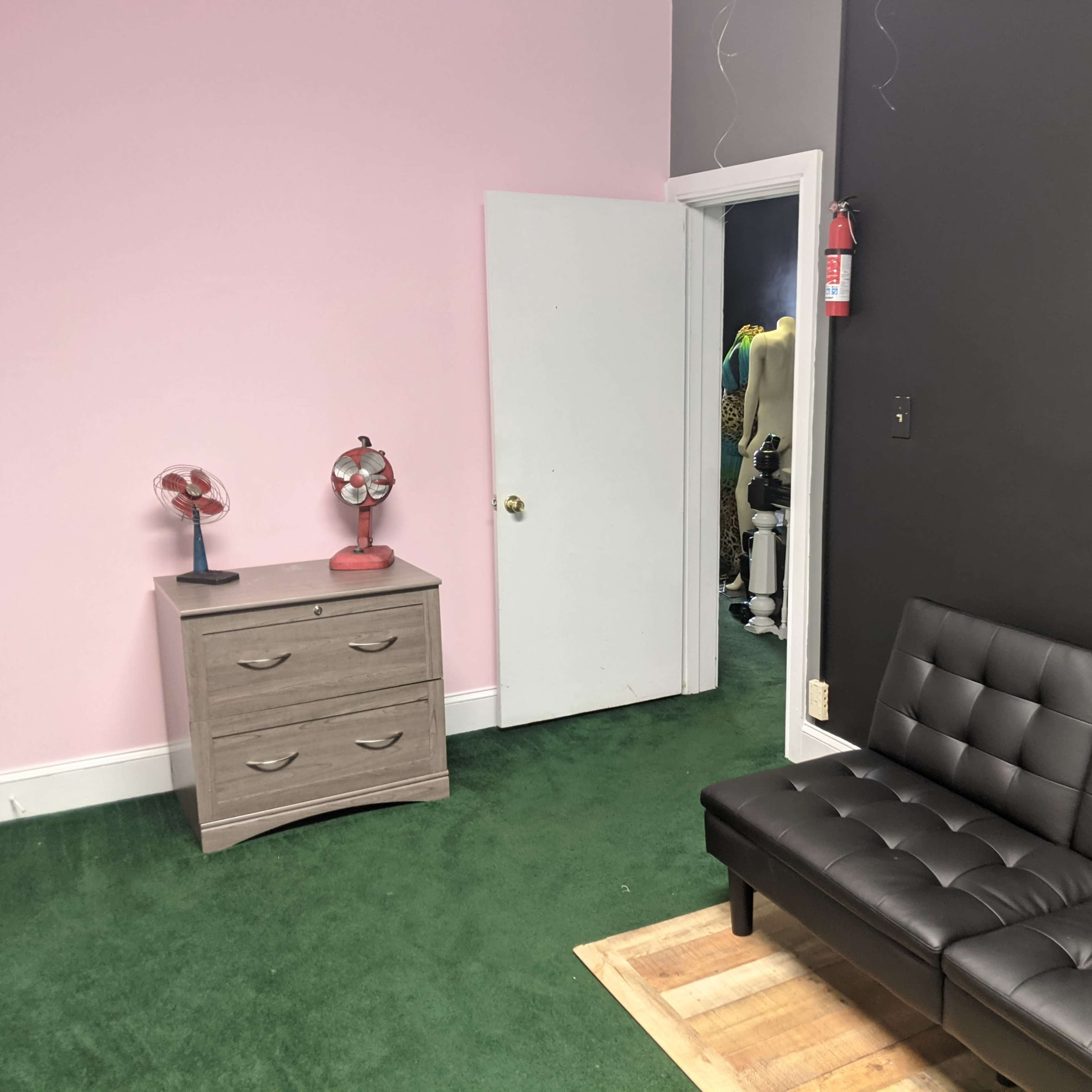 A room with a pink wall, a black wall, a gray dresser, a black couch, and a door leading to another area.