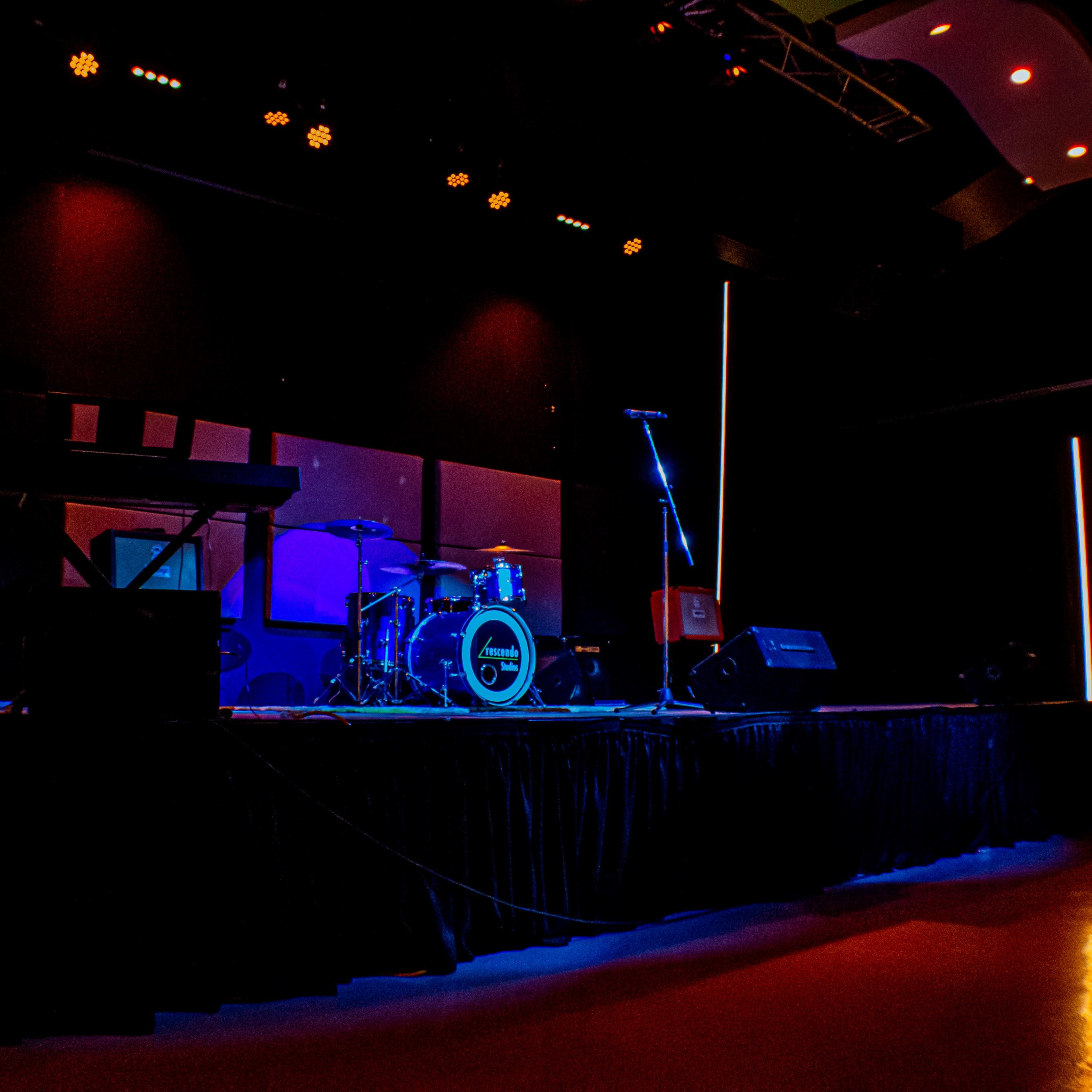 The stage is set for a musical performance, featuring a drum kit, microphone stand, and amplifiers, illuminated by colored lights.