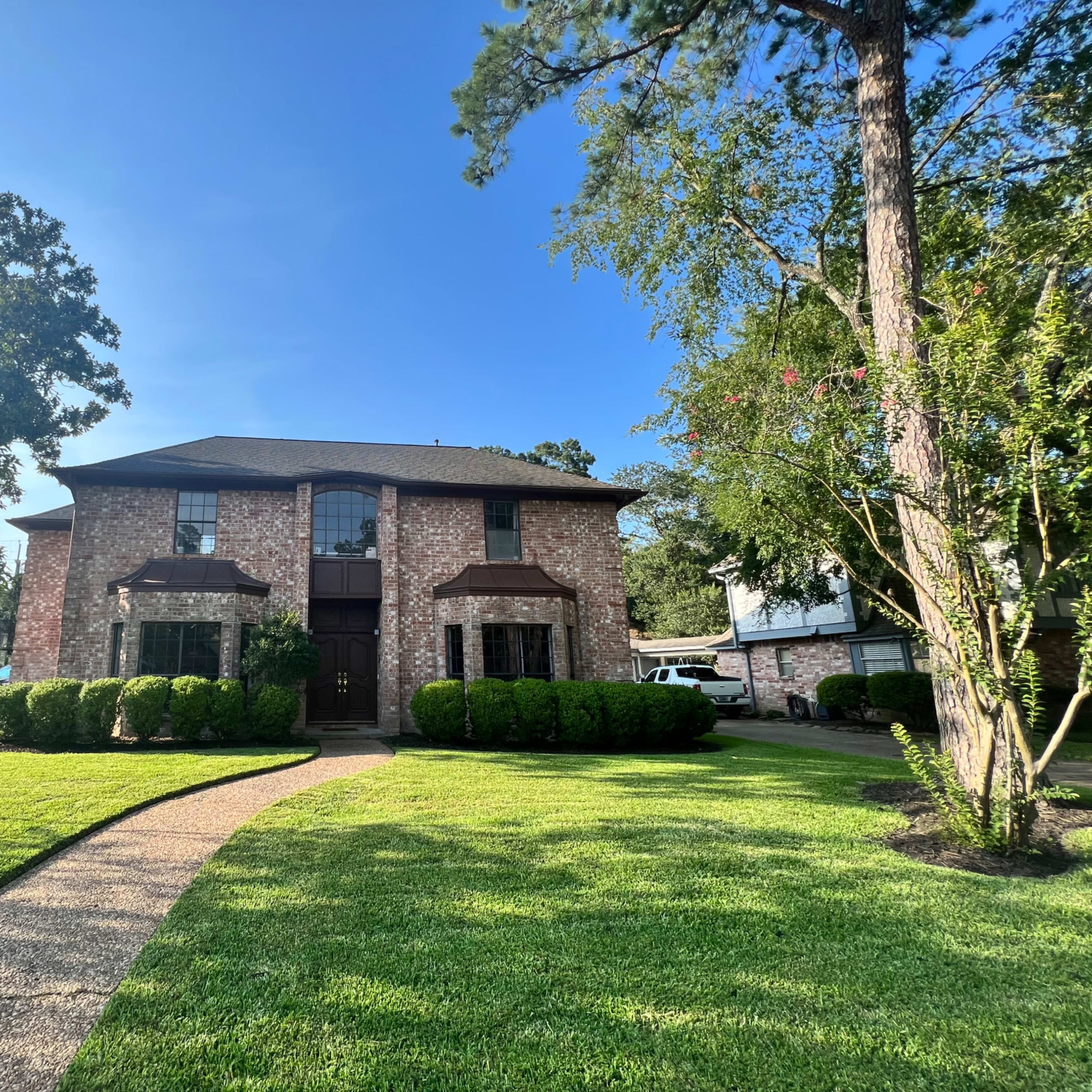 A two-story brick house with flat-roofed entryways is surrounded by manicured lawns and tall trees.