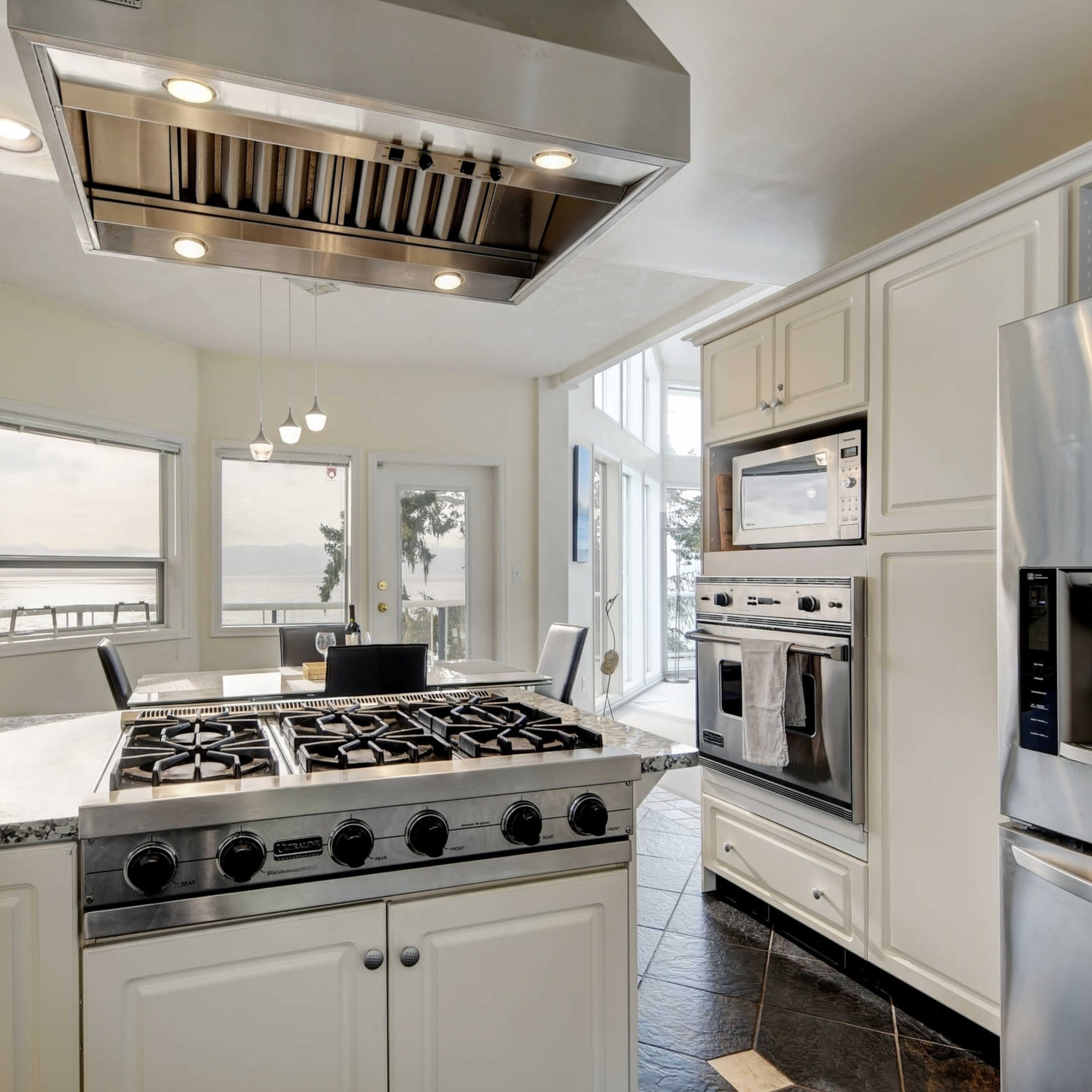 A modern kitchen features stainless steel appliances, a large gas range, and a view of a waterfront through tall windows.