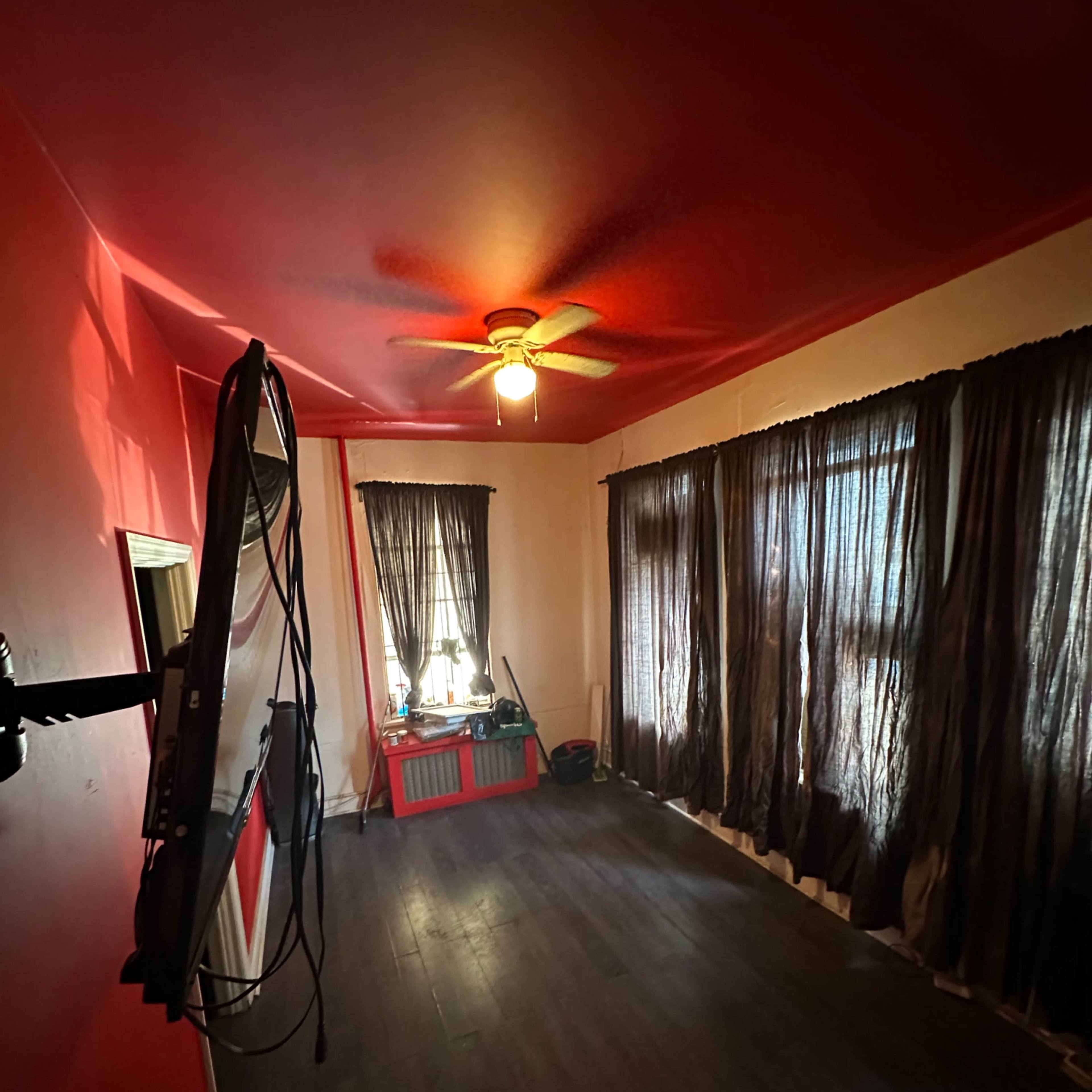 A room with red walls, a ceiling fan, a mounted television, and long dark curtains over windows.