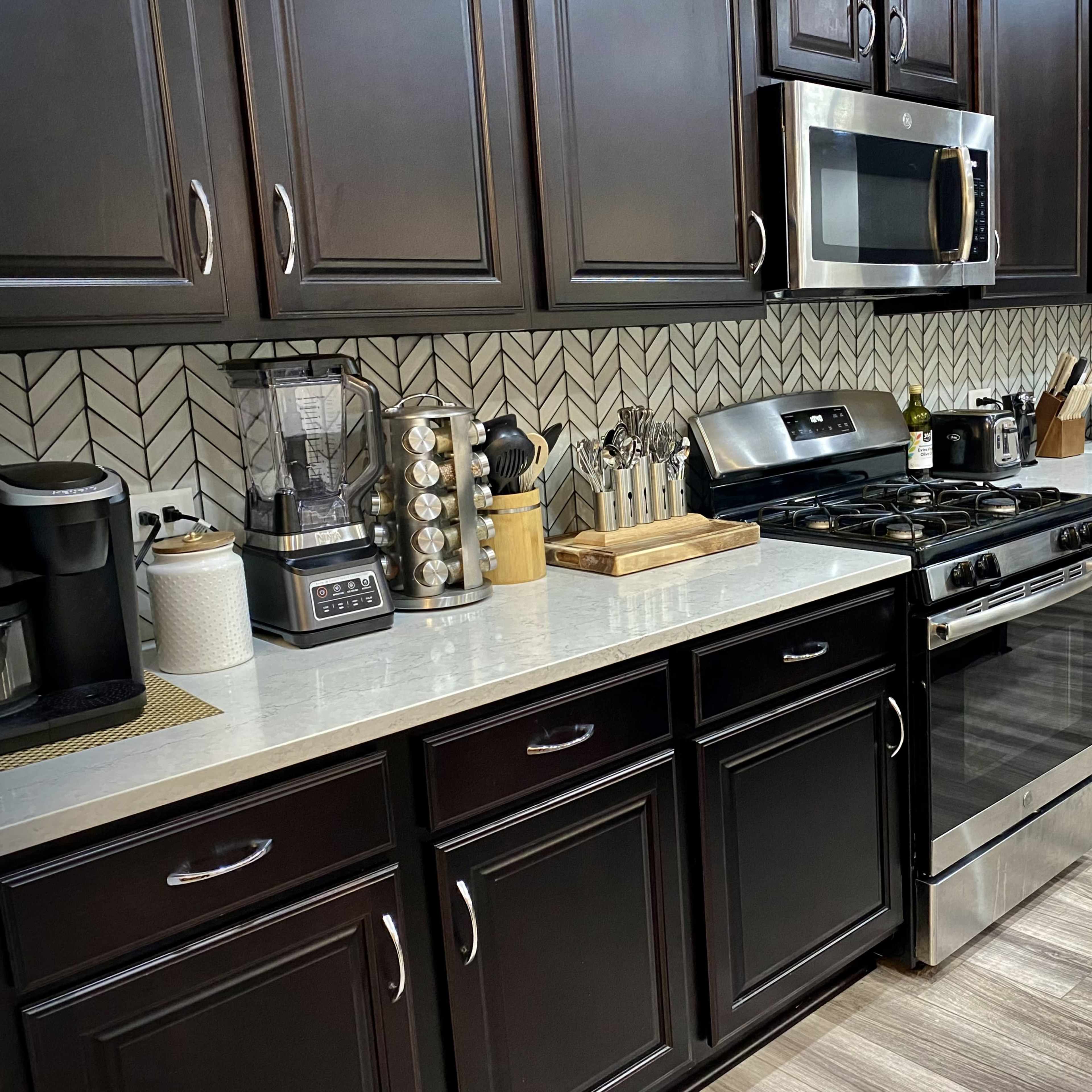 A modern kitchen features dark cabinetry, stainless steel appliances, and a light countertop with various kitchen tools and appliances on display.