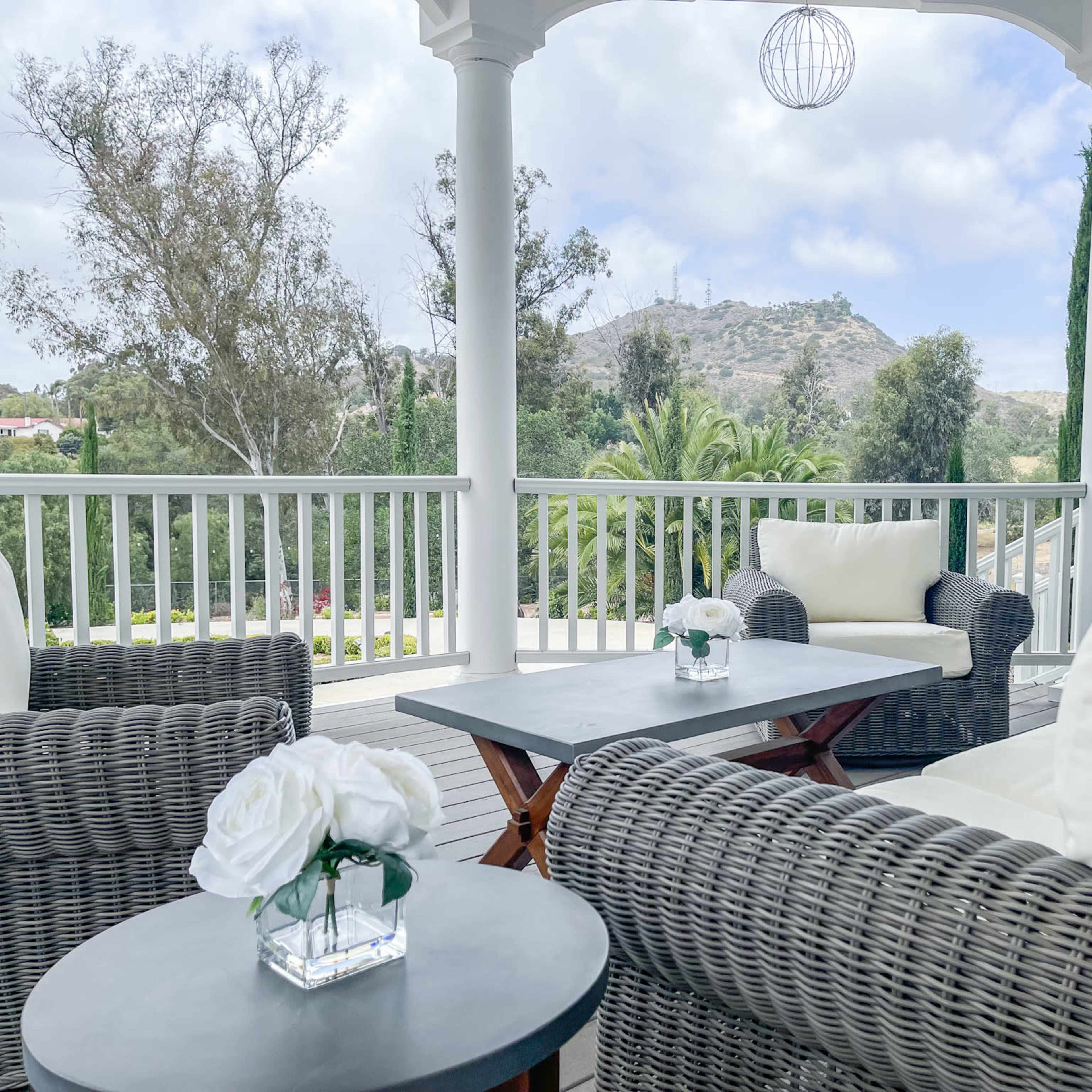 A covered outdoor patio with wicker furniture, a small table with a floral centerpiece, and a view of a hillside in the background.