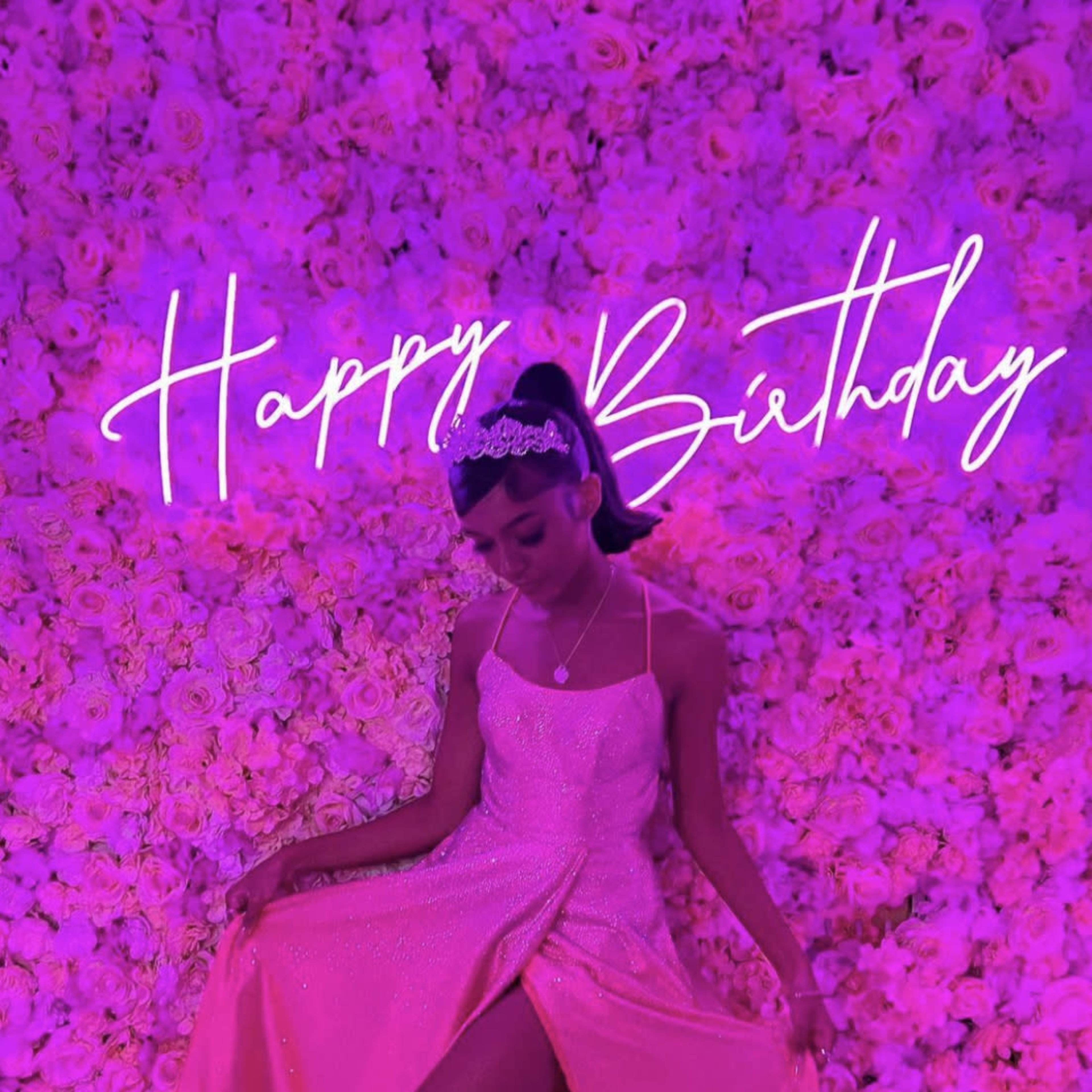 A person in a pink dress poses in front of a wall covered with artificial flowers, with "Happy Birthday" written in bright letters above.
