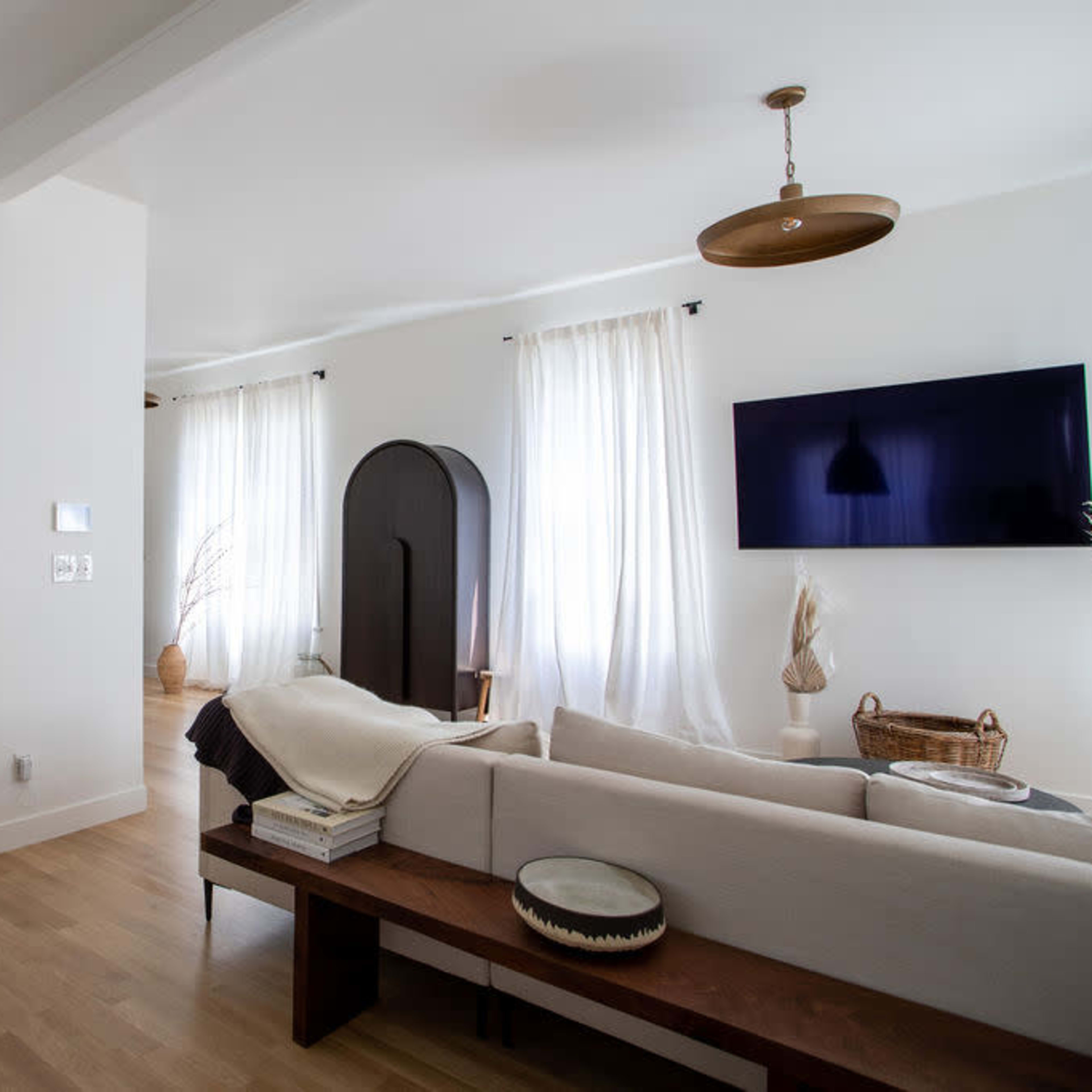 A modern living room features a colorful abstract painting, a neutral sofa with a throw, a wooden bench, and a television mounted on the wall.