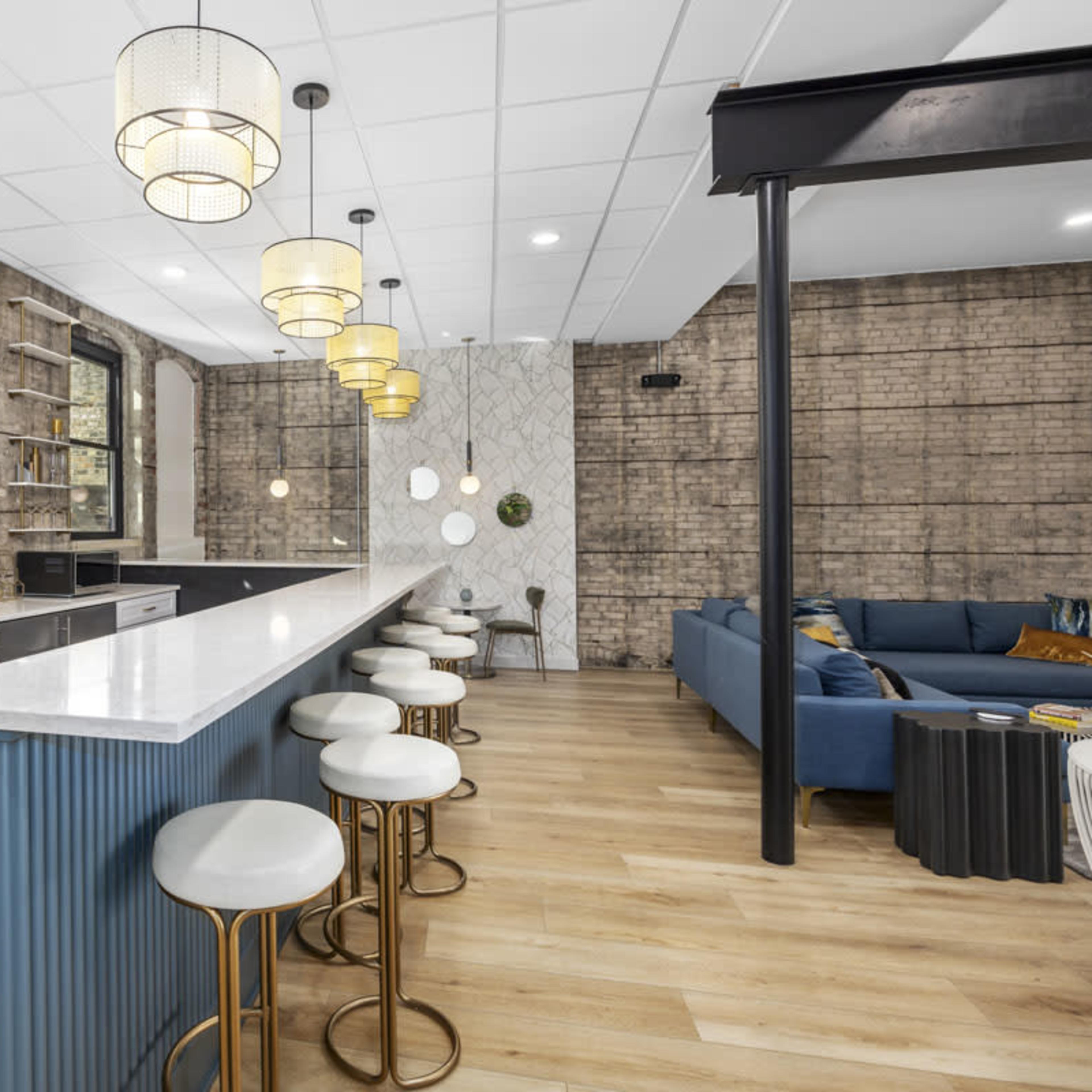 A modern kitchen features a long, white countertop with high chairs, set against a backdrop of exposed brick walls and stylish pendant lighting.