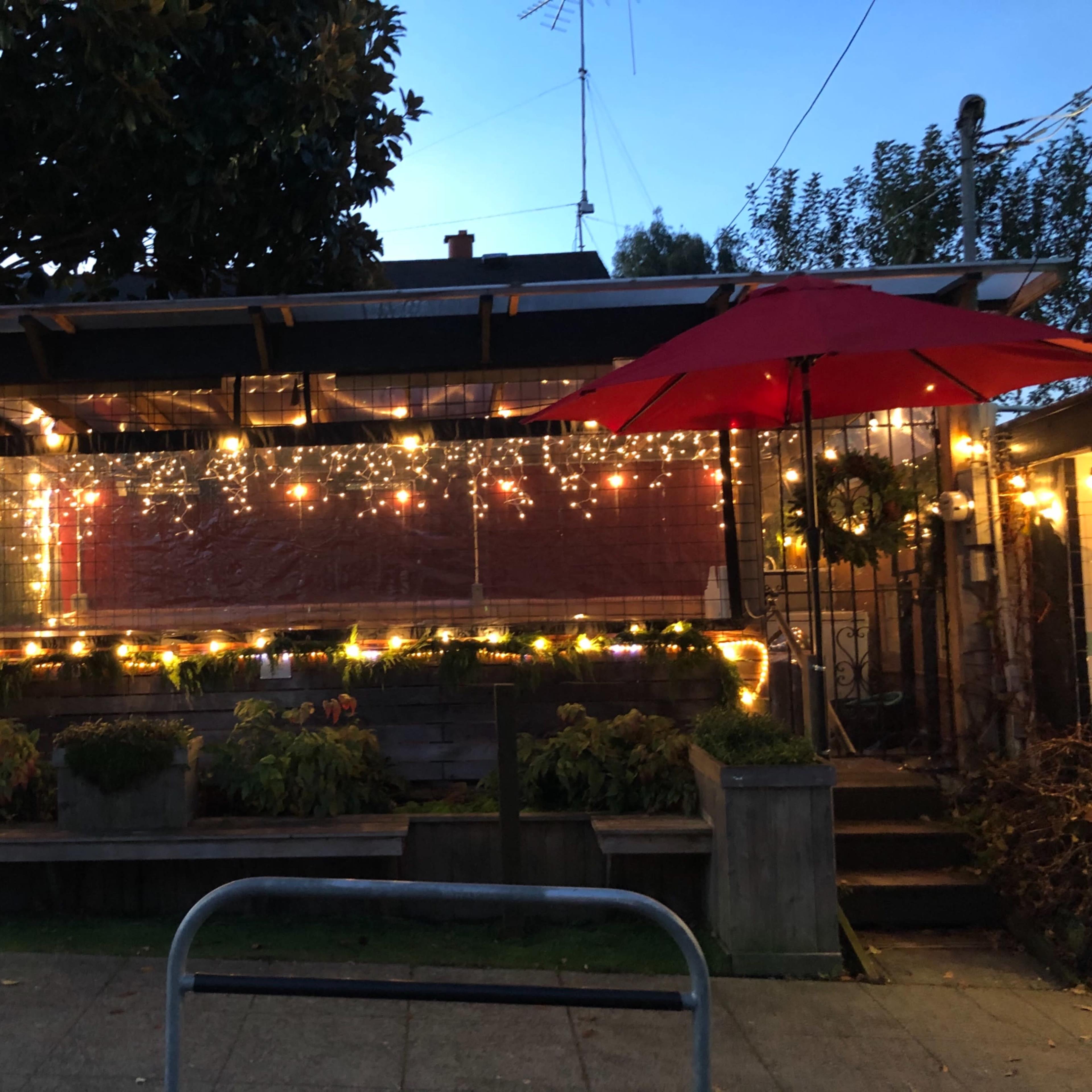 The exterior of a building features outdoor seating under red umbrellas and is adorned with string lights, creating a cozy atmosphere.