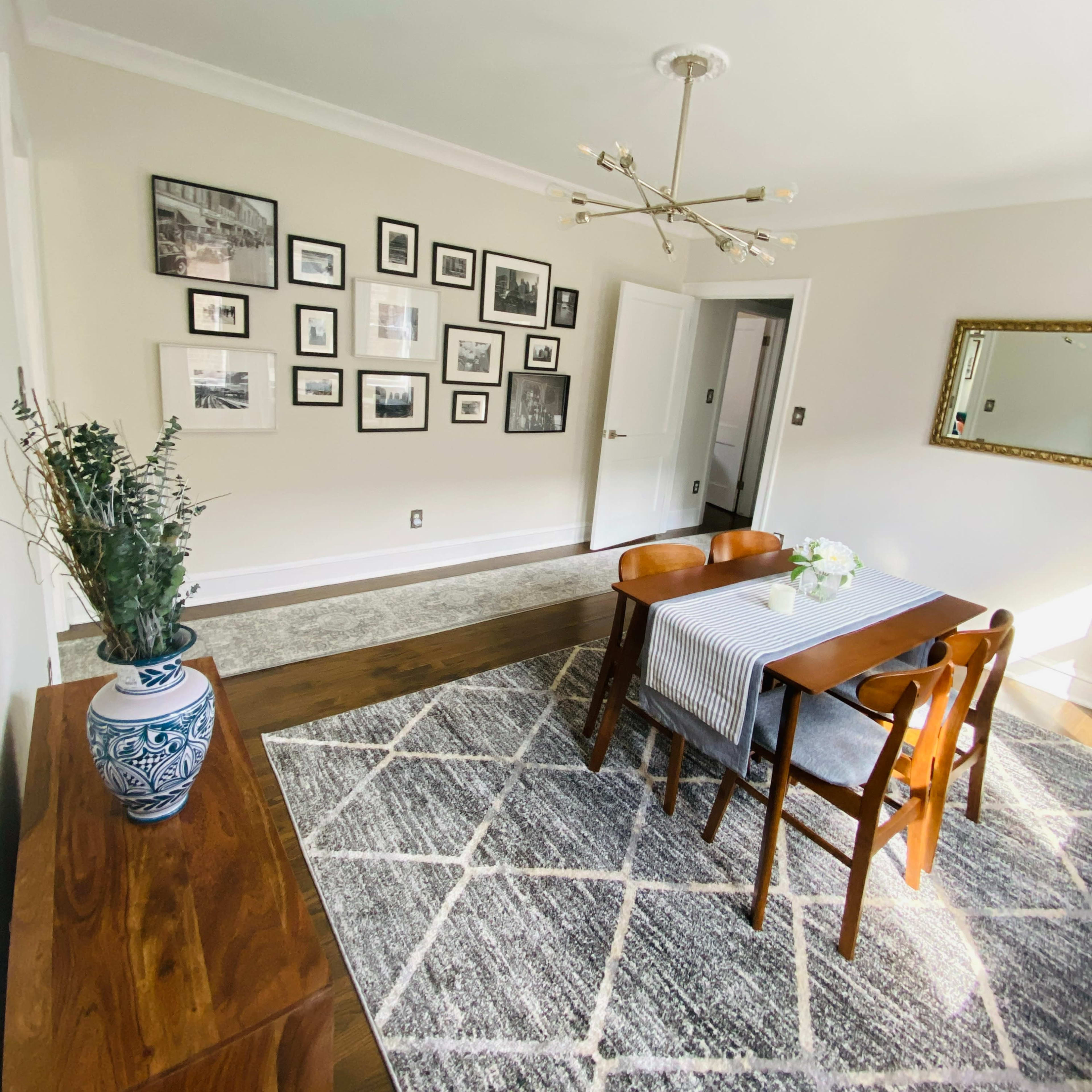A dining area features a wooden table with chairs, a patterned rug, and a wall adorned with framed pictures.