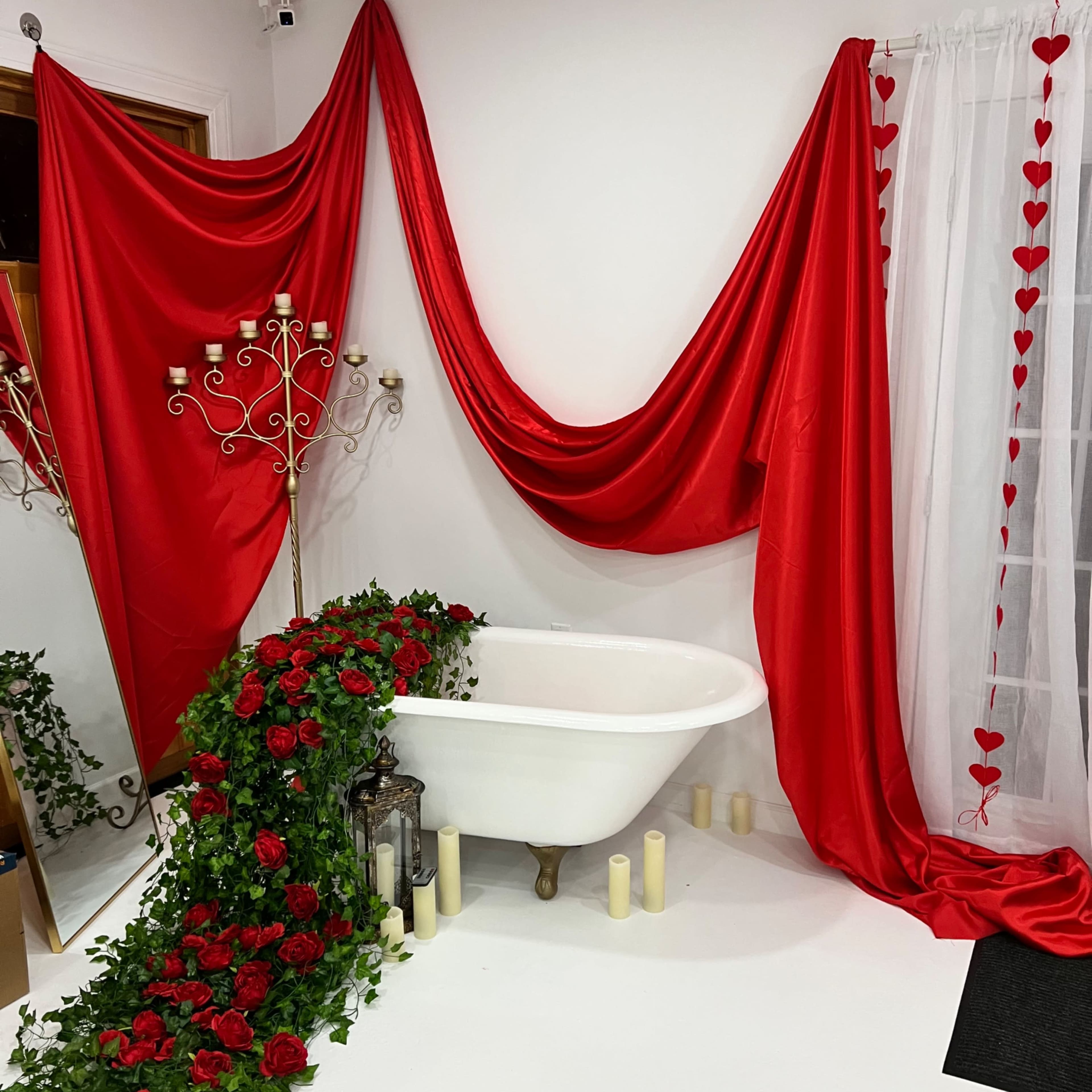 A white bathtub is adorned with red fabric and greenery, surrounded by candles and a decorative lantern.