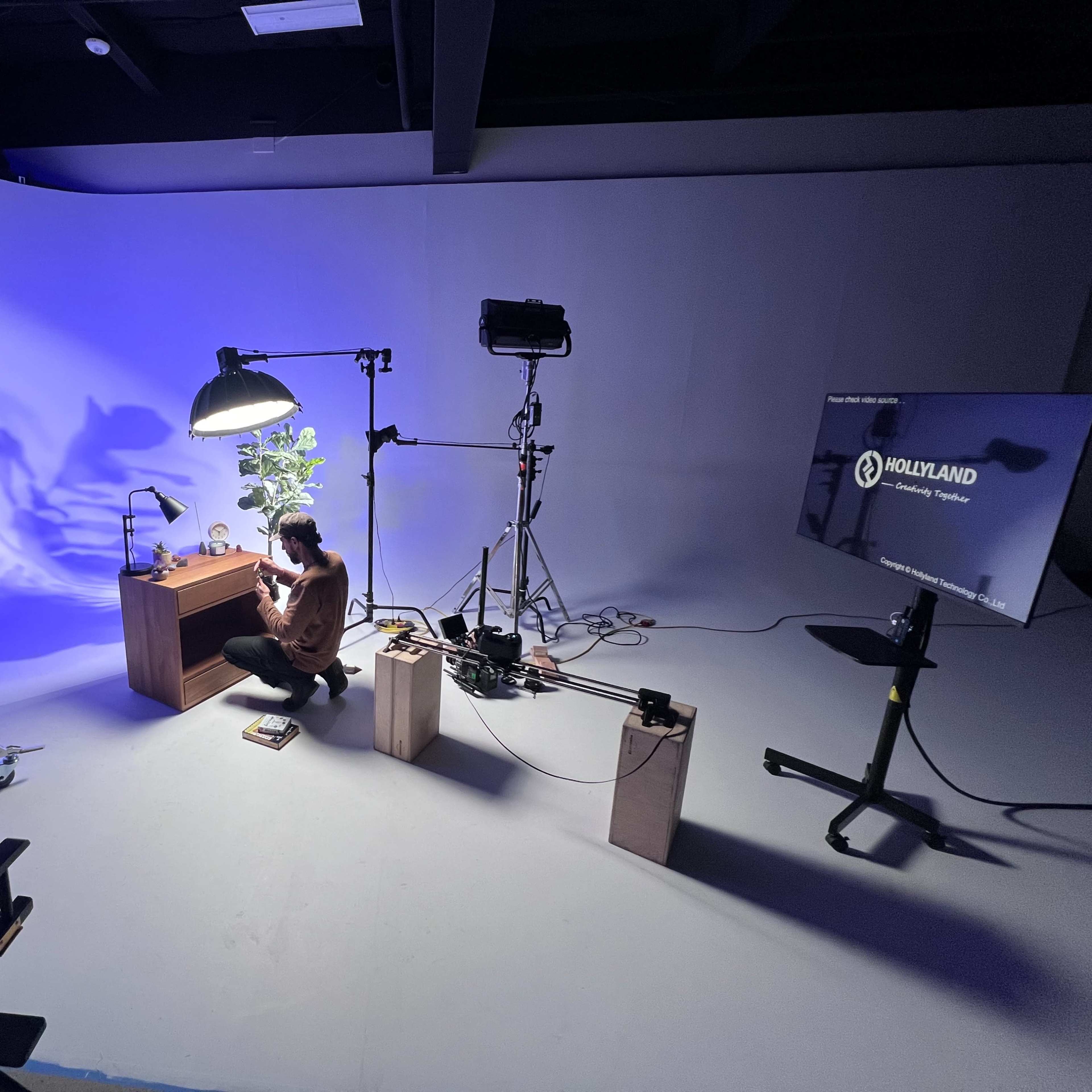 A person is setting up a scene in a studio with a desk, lighting equipment, and a plant, while a monitor displays content in the background.