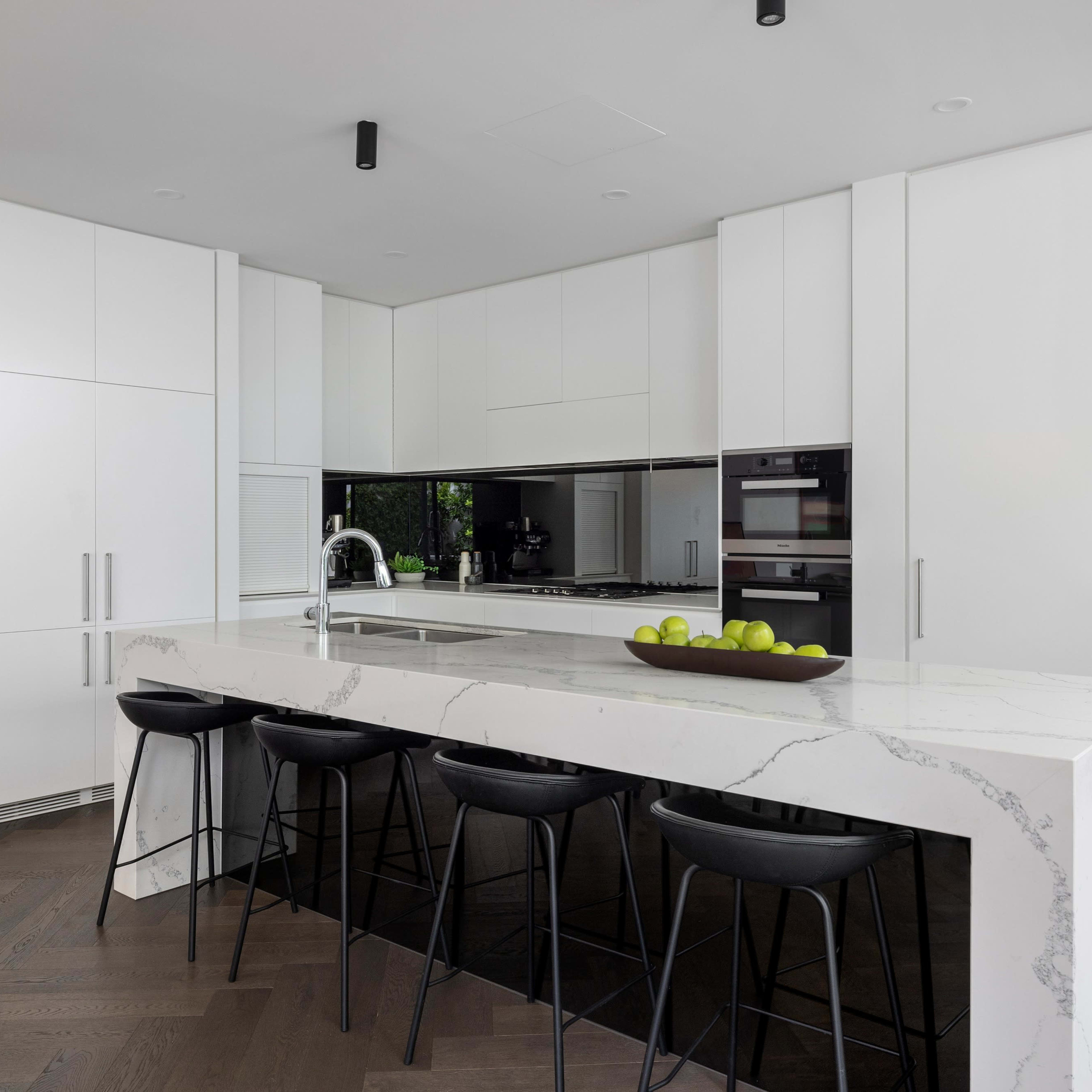 A modern kitchen featuring white cabinetry, a marble-topped island with four black stools, and a green plant on a shelf.