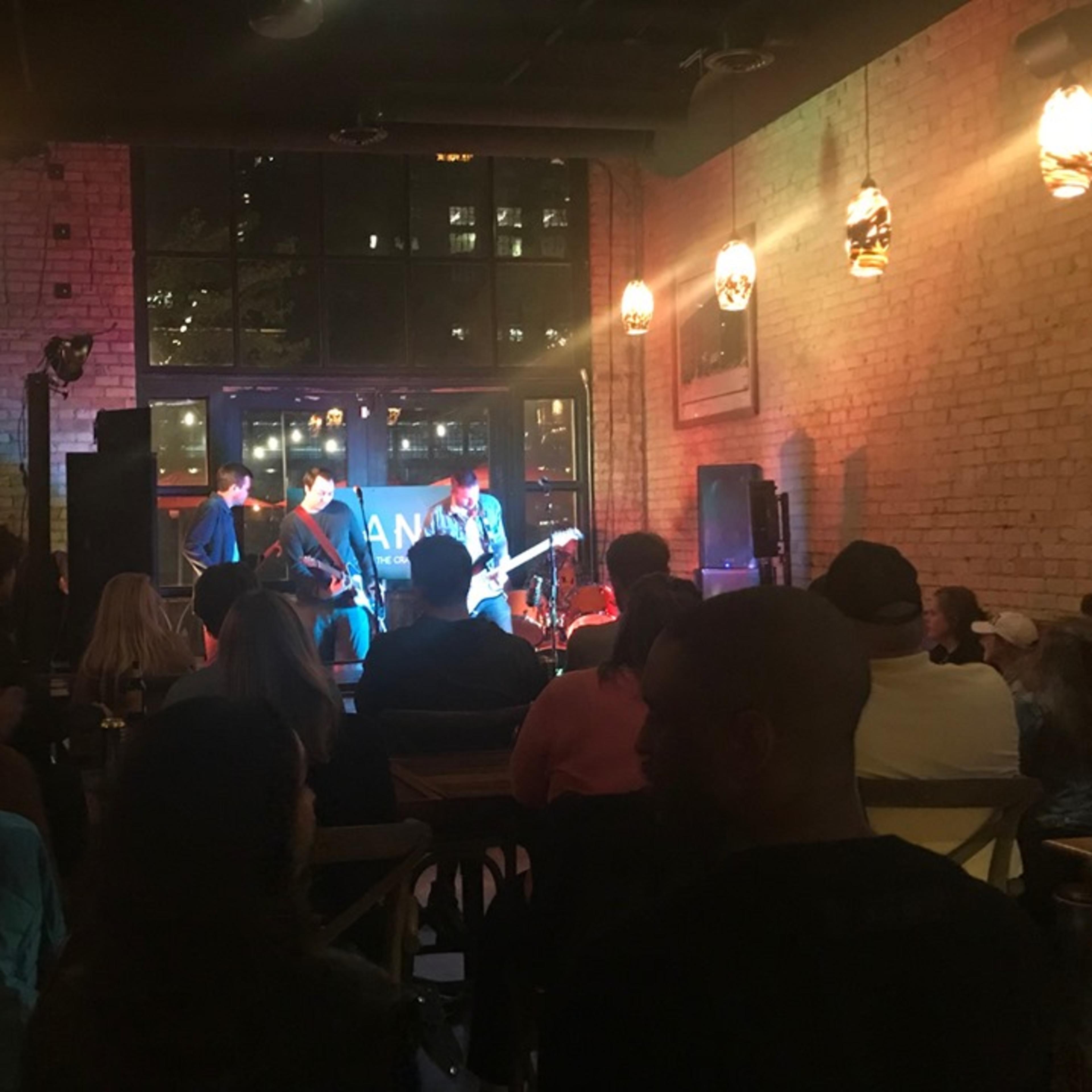 A small crowd watches a live band perform inside a dimly lit venue with large windows.