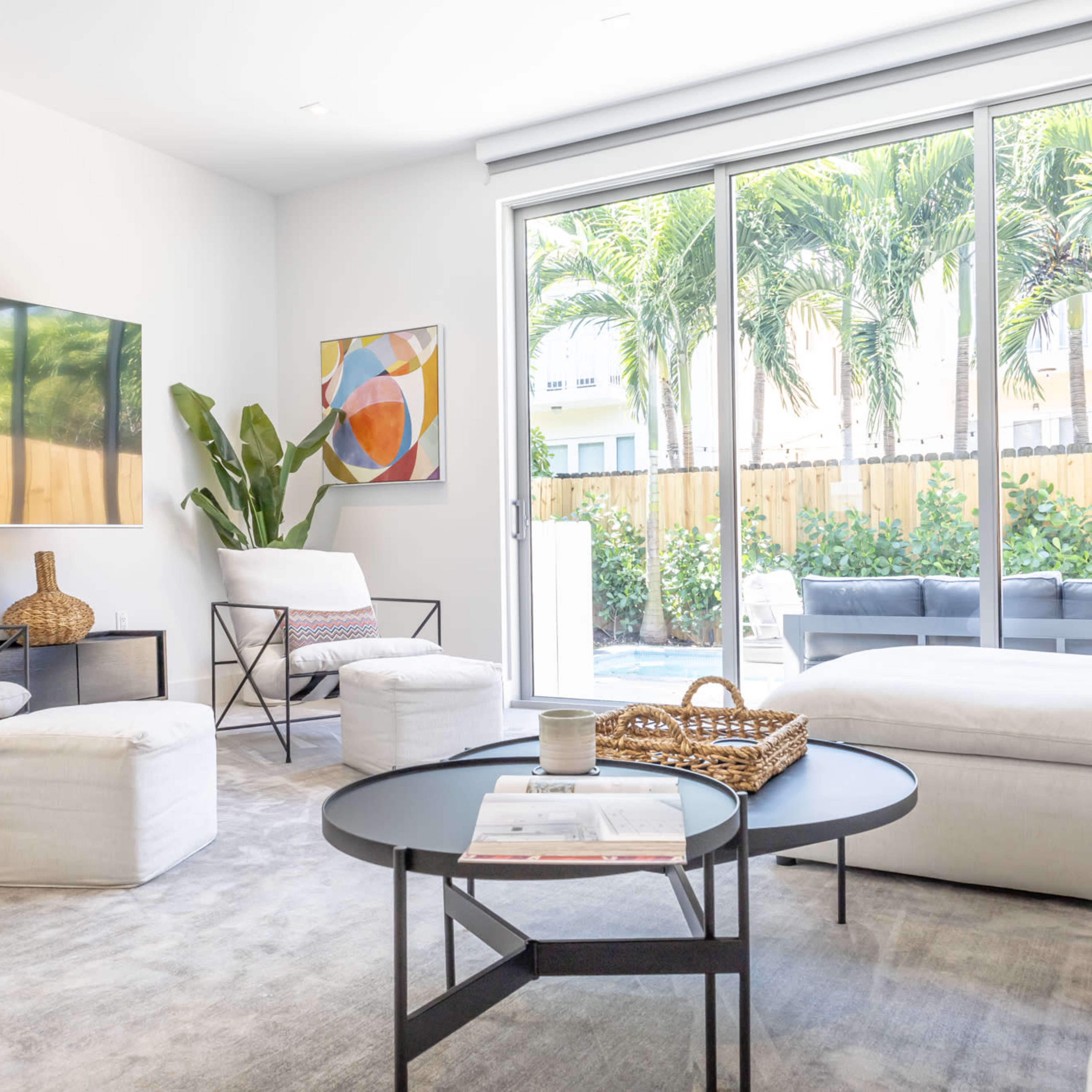 A modern living room with a large window overlooking a tropical garden, featuring light-colored furniture and a coffee table in the center.