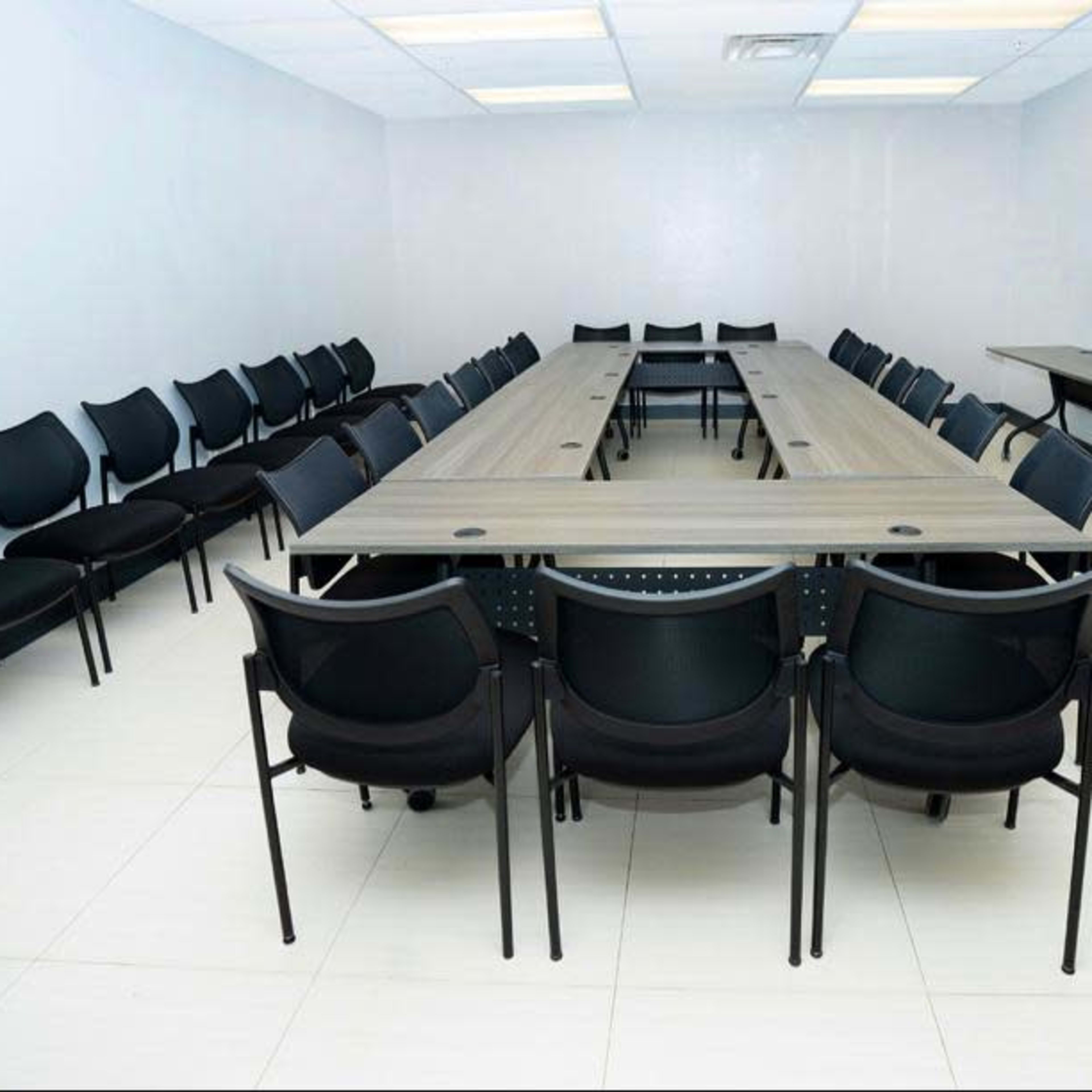 A rectangular conference room with a long table surrounded by black chairs and a television mounted on a stand against one wall.