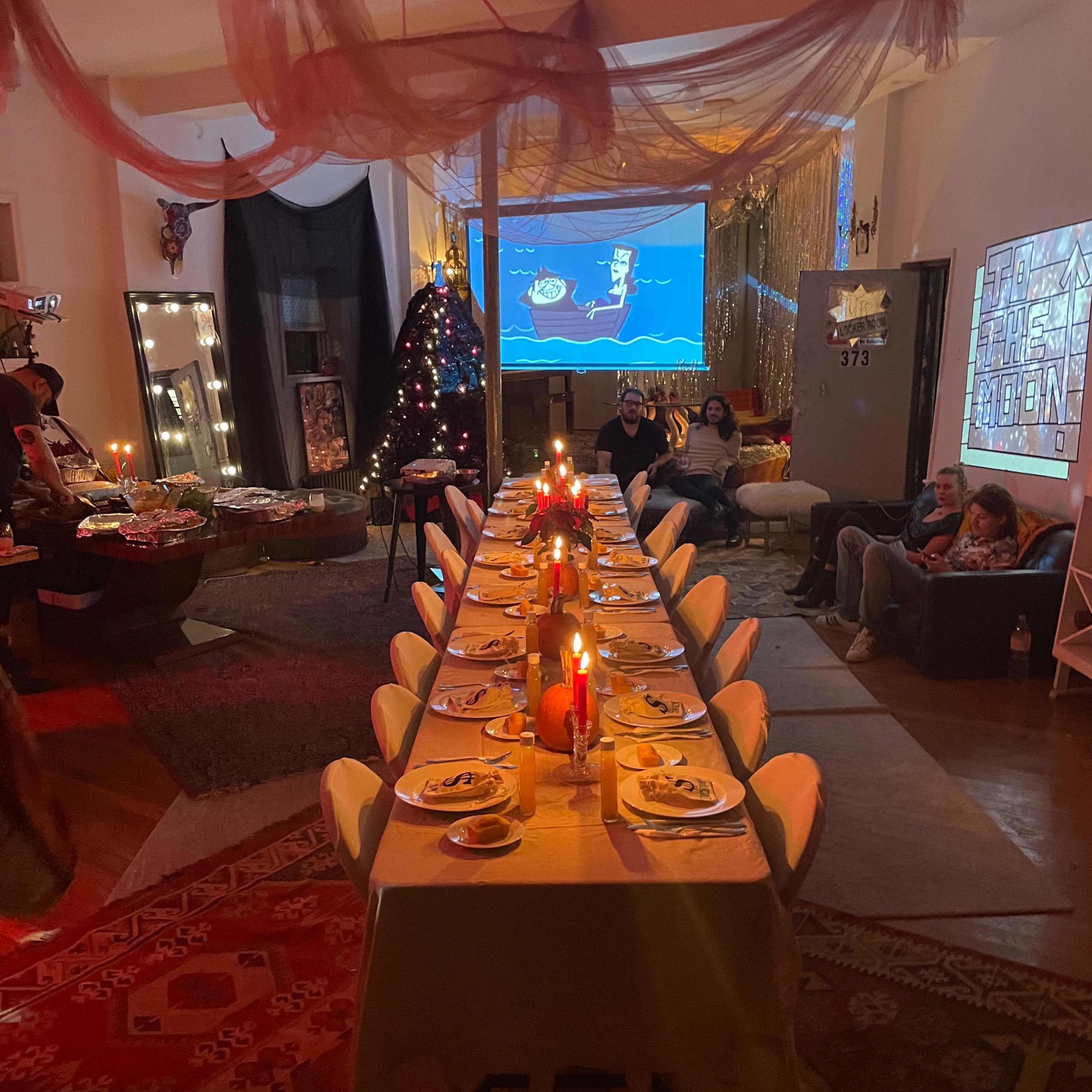 A long dining table set for a gathering, decorated with candles and plates, in a festively arranged room featuring a Christmas tree and a projection on the wall.