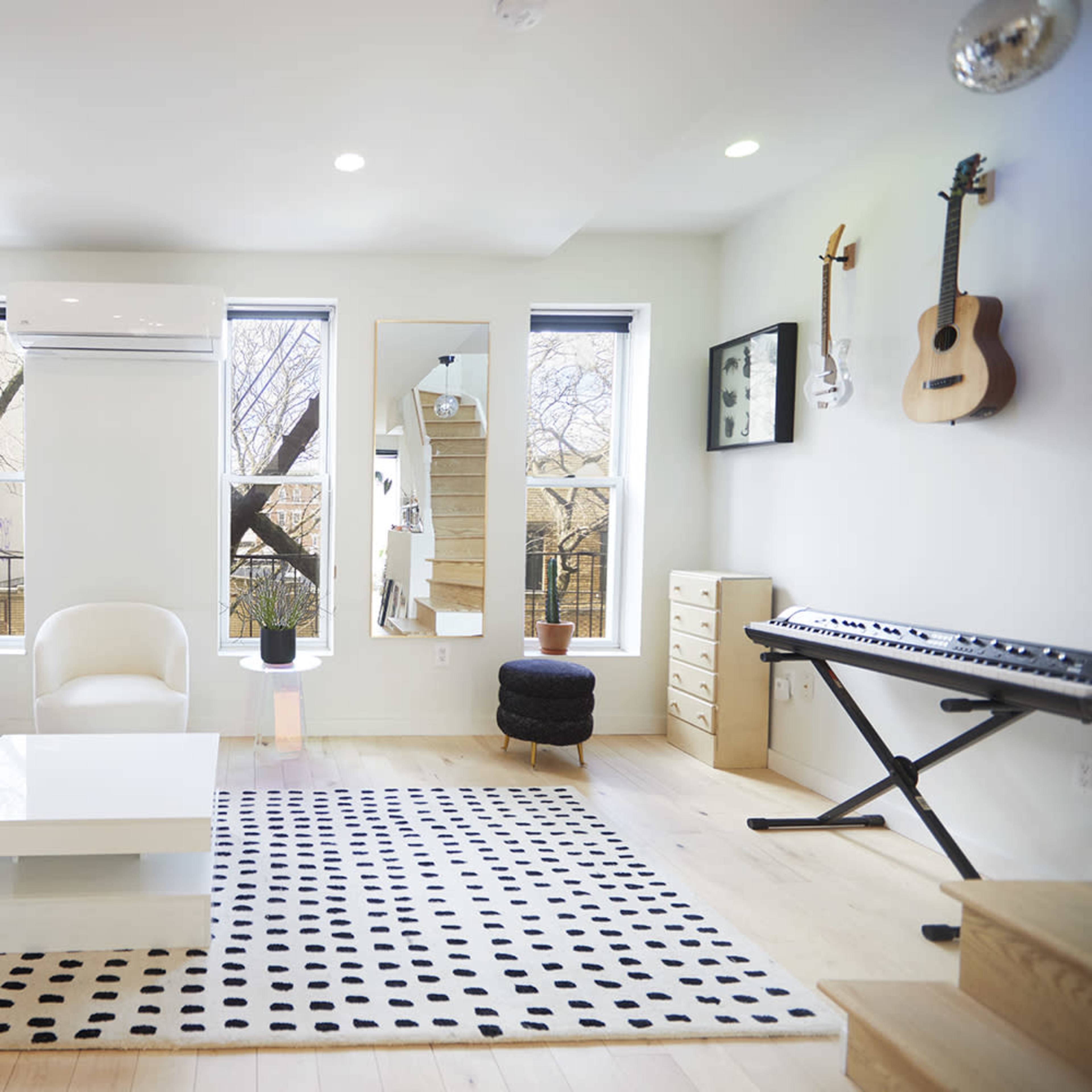 A modern living room features a gray sofa, a white armchair, a black and white polka dot rug, and a keyboard, with large windows providing natural light.