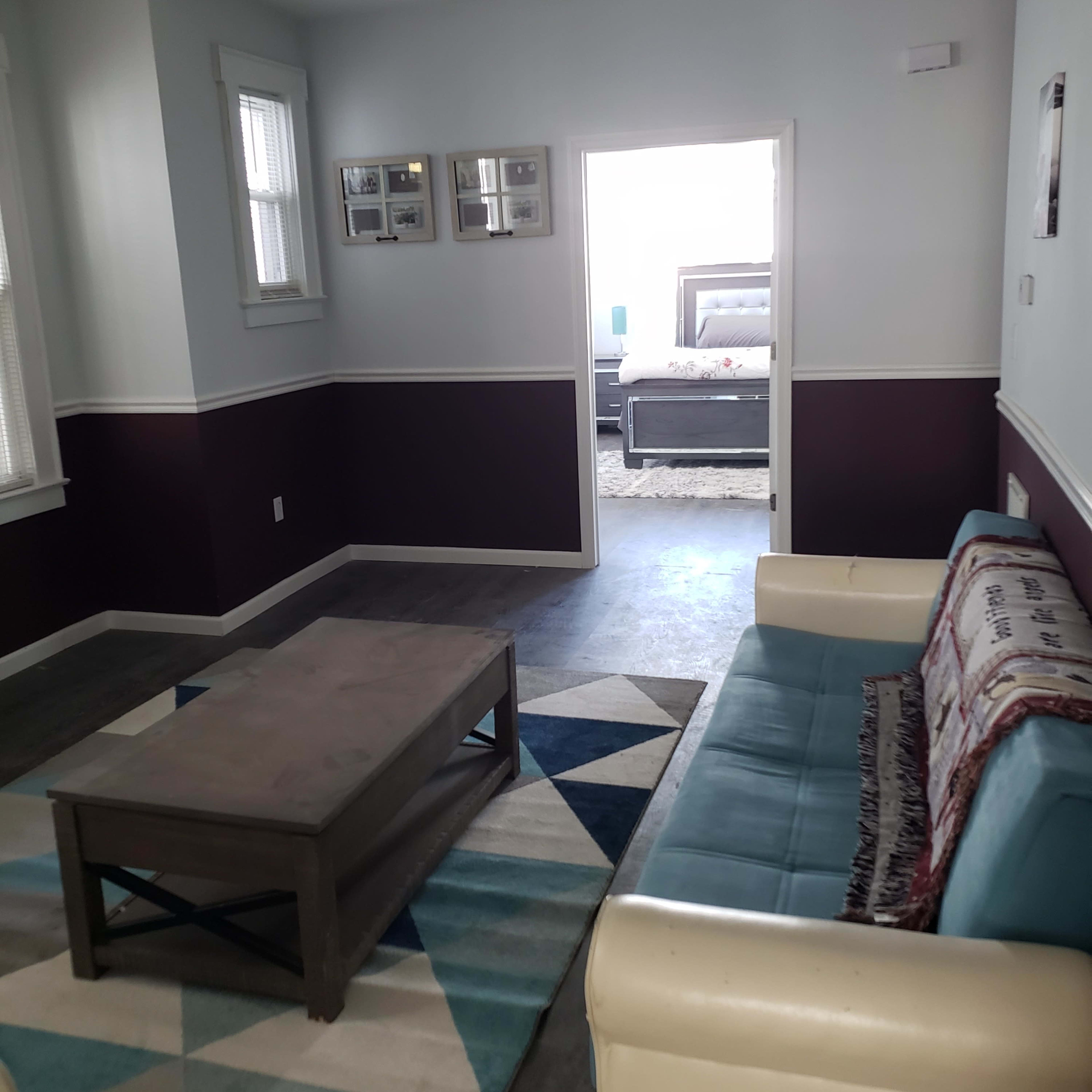A living room featuring a teal sofa, a coffee table, and a geometric rug, with a doorway leading to a bedroom in the background.
