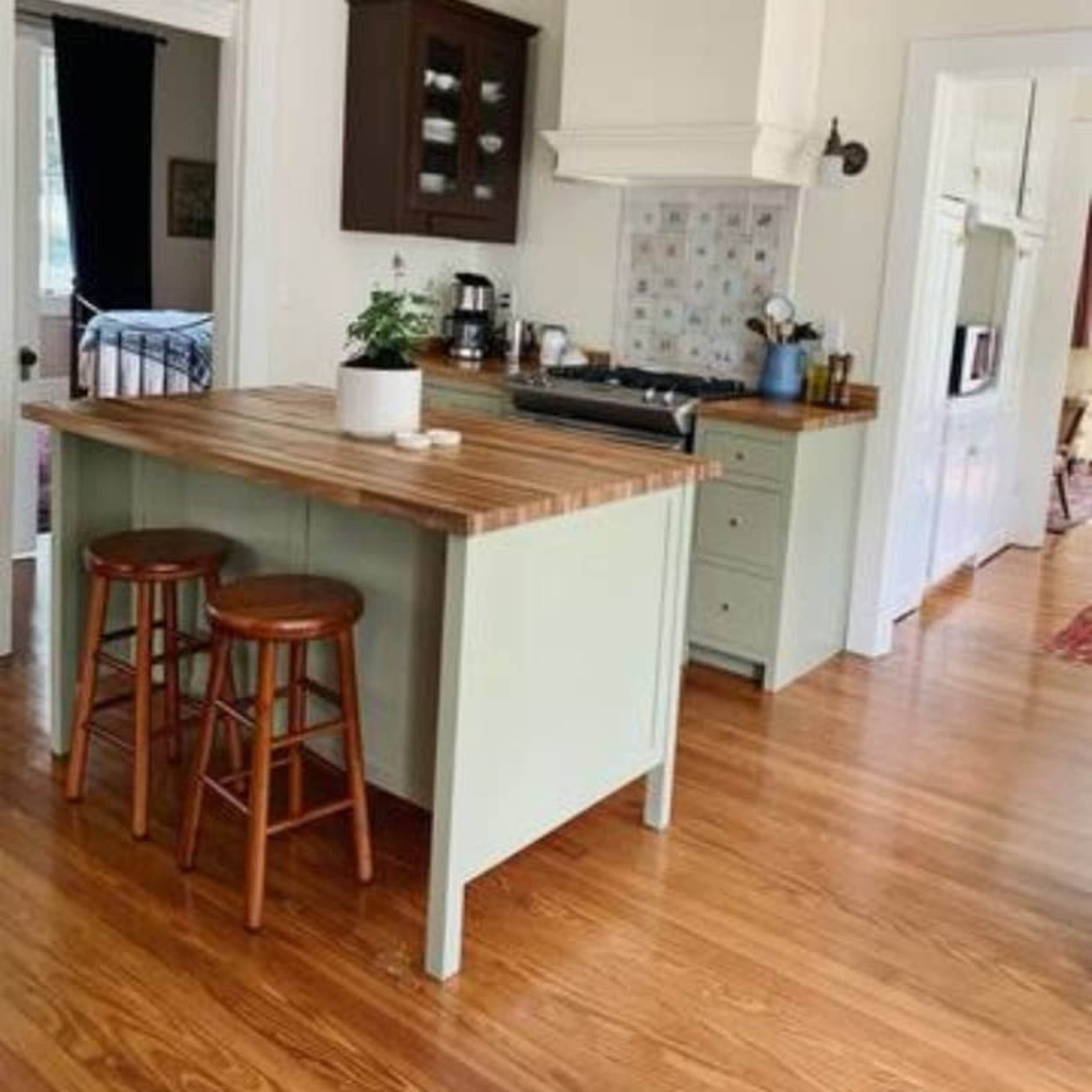 A light-colored kitchen features a wooden island with two stools, a stove, and cabinetry, with a doorway leading to another room in the background.
