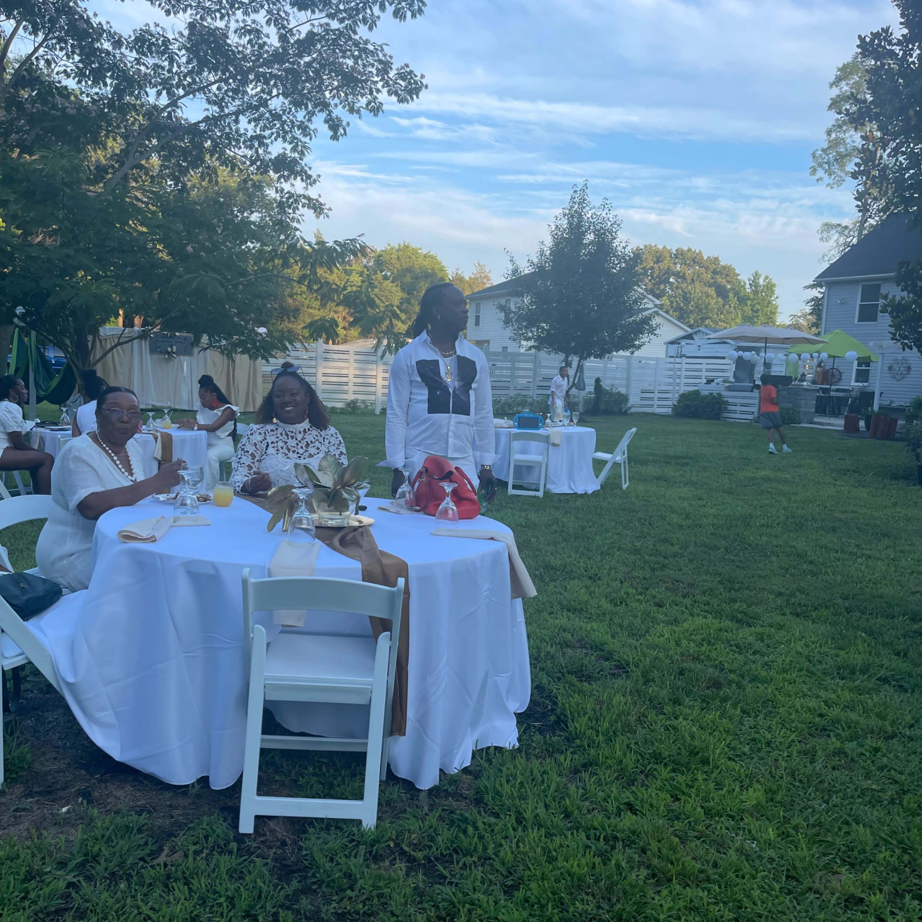 A group of people is seated at tables in a grassy outdoor area during a gathering, while another person stands nearby.