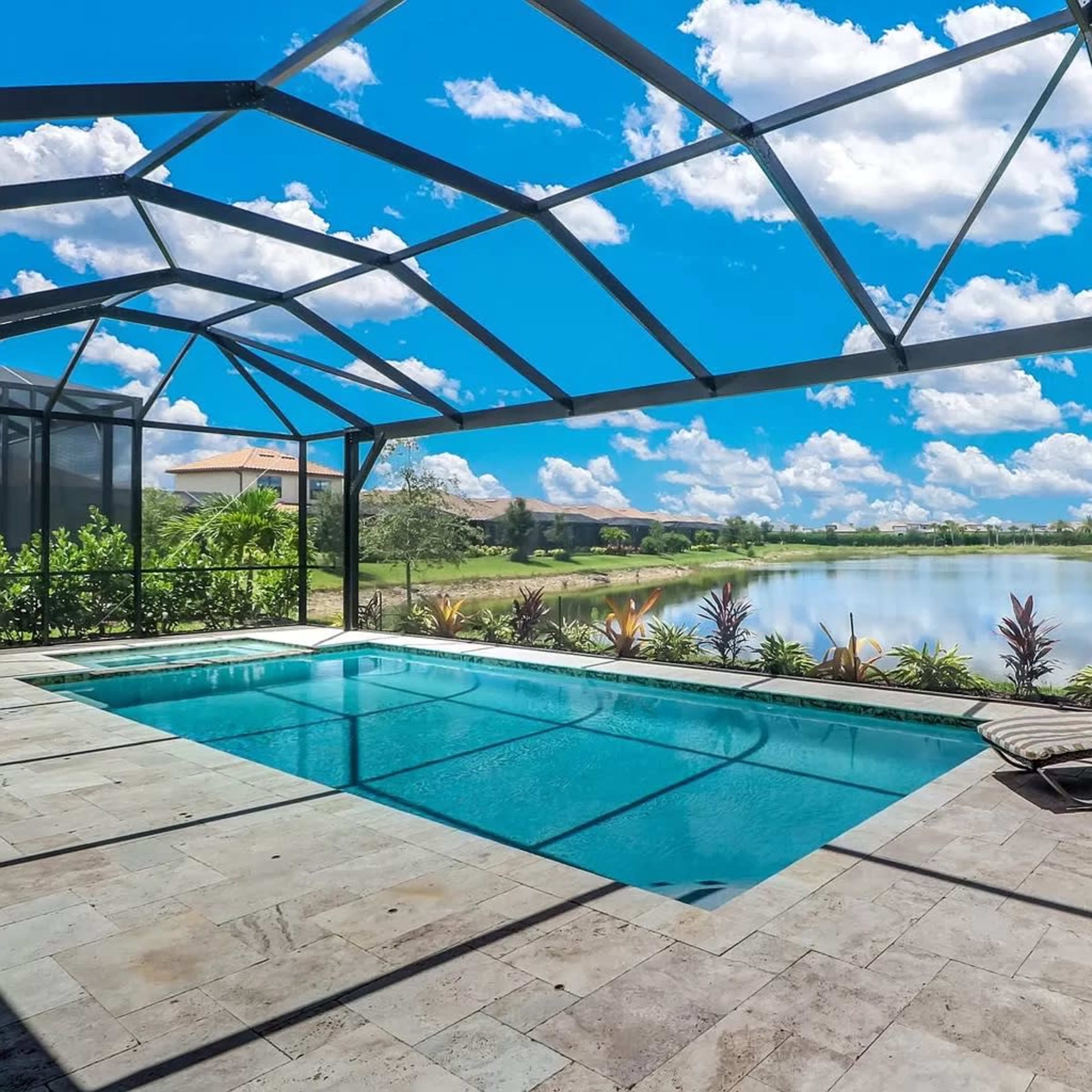 A screened pool area with two lounge chairs overlooks a calm lake surrounded by lush greenery under a bright sky.
