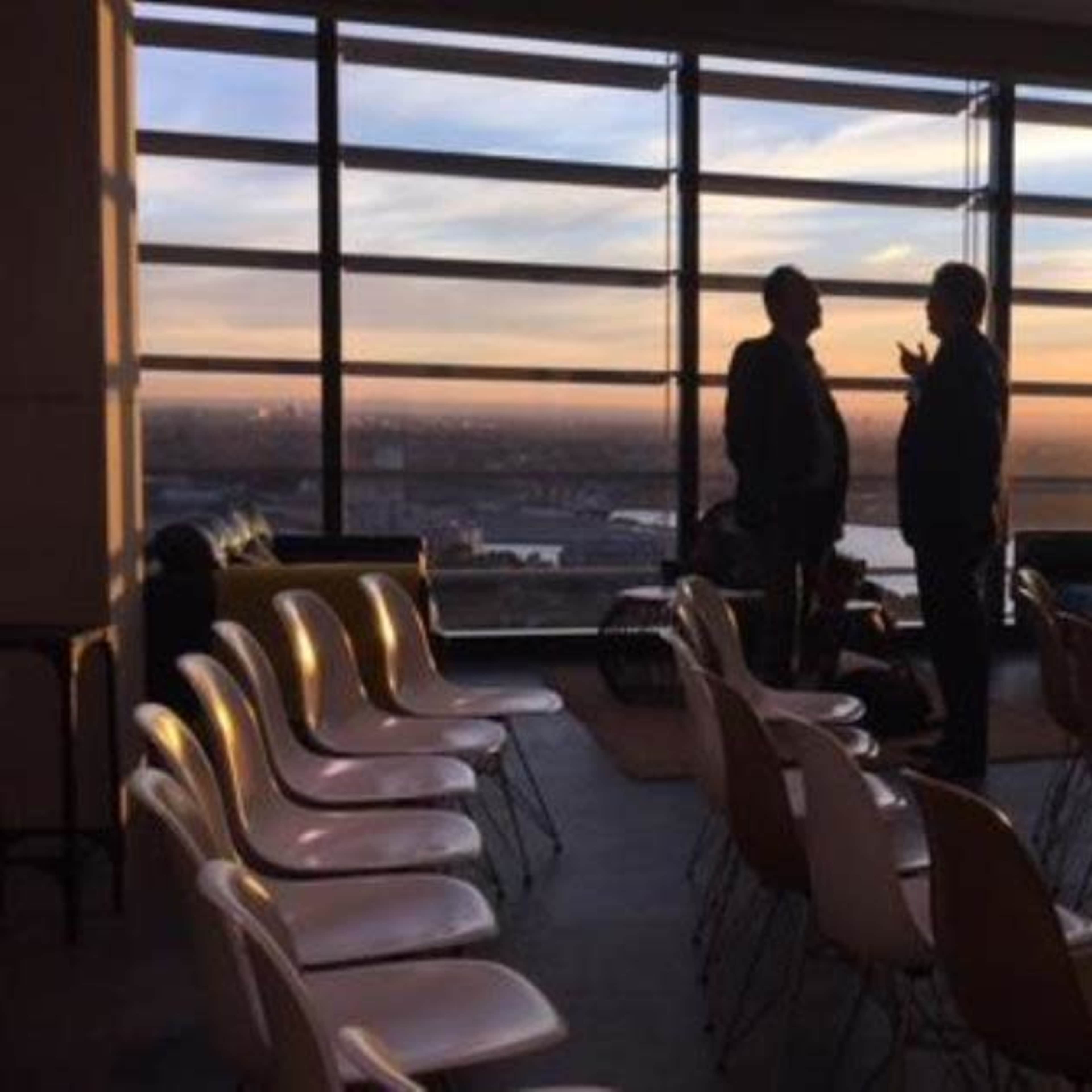 Two people stand and converse in a room with rows of empty chairs and large windows displaying a sunset view.
