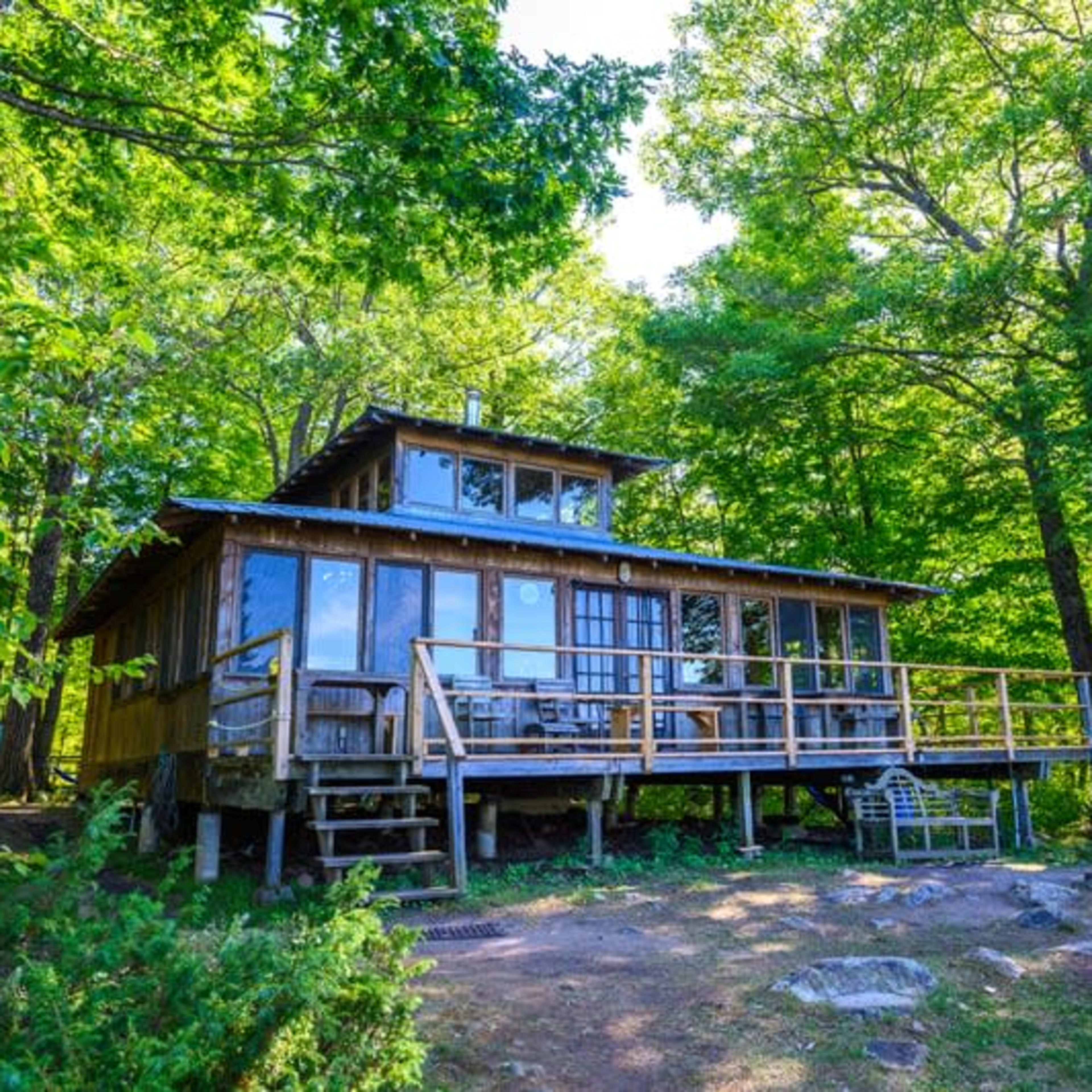 A wooden cabin with large windows is situated on a hillside surrounded by green trees.