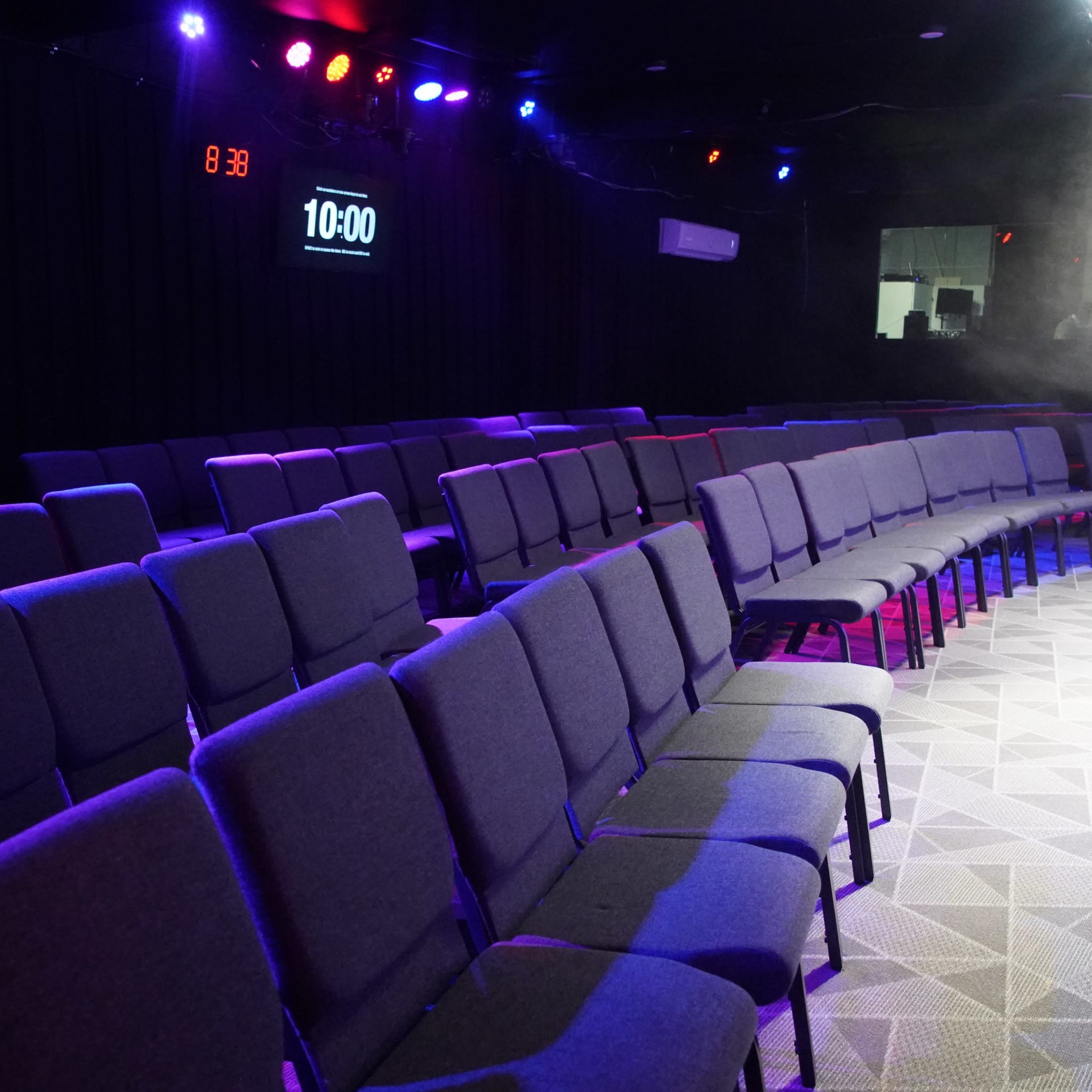 A row of gray chairs is arranged in a semi-circular layout in a dimly lit auditorium with colored lights and a time display on the wall.