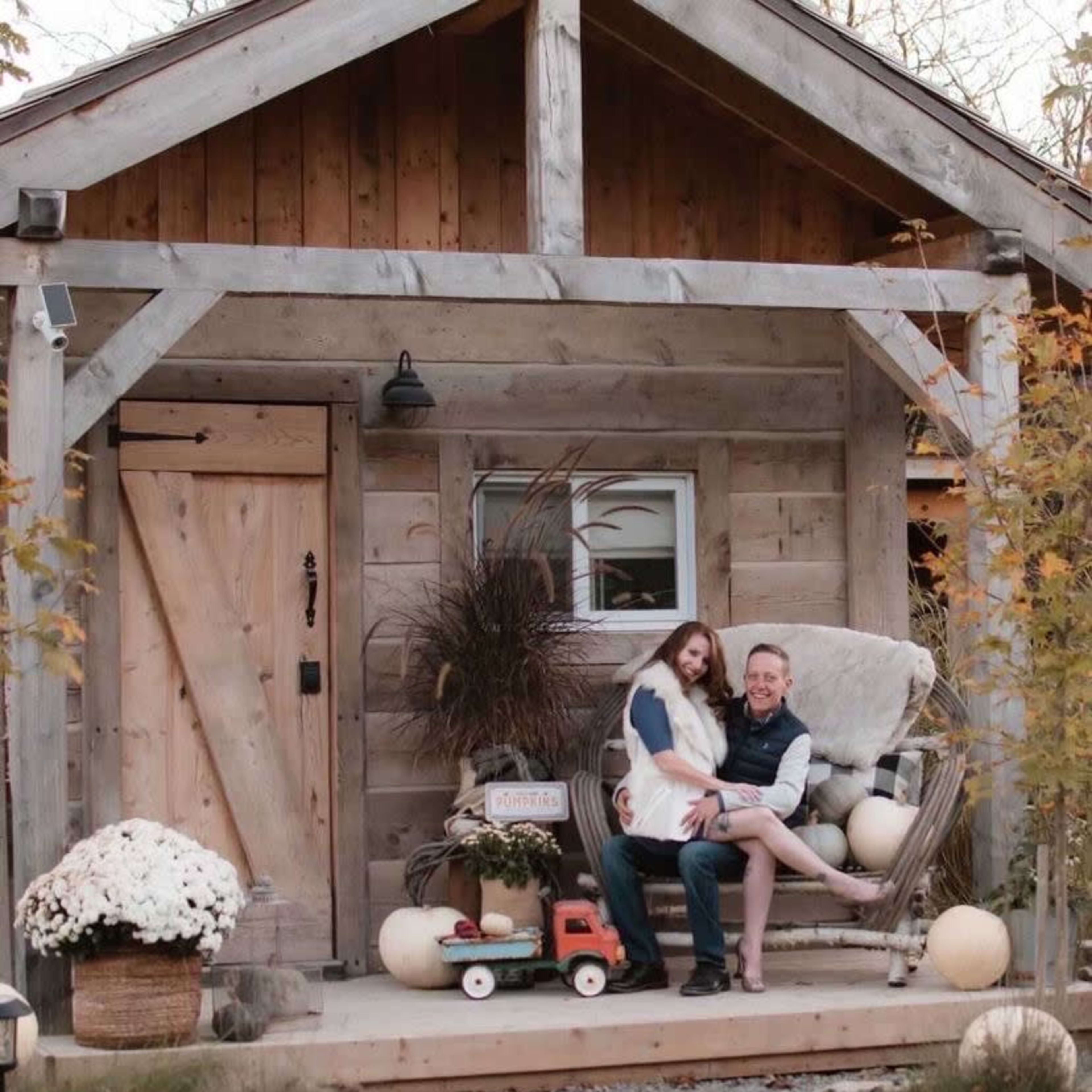 A couple sits on a rocking chair on the porch of a rustic wooden cabin, surrounded by decorative pumpkins and flowers.
