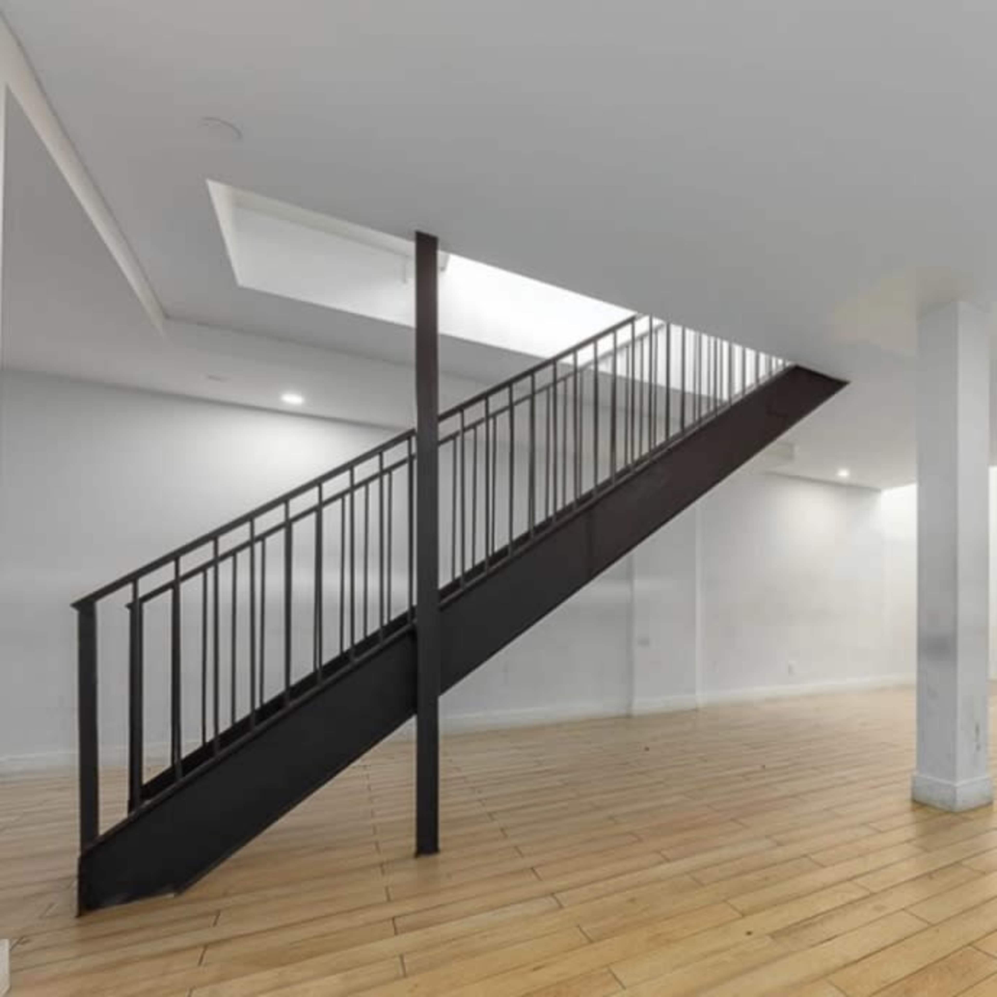 An empty room with wooden flooring and a staircase on the left leading to an upper floor, illuminated by recessed lighting.