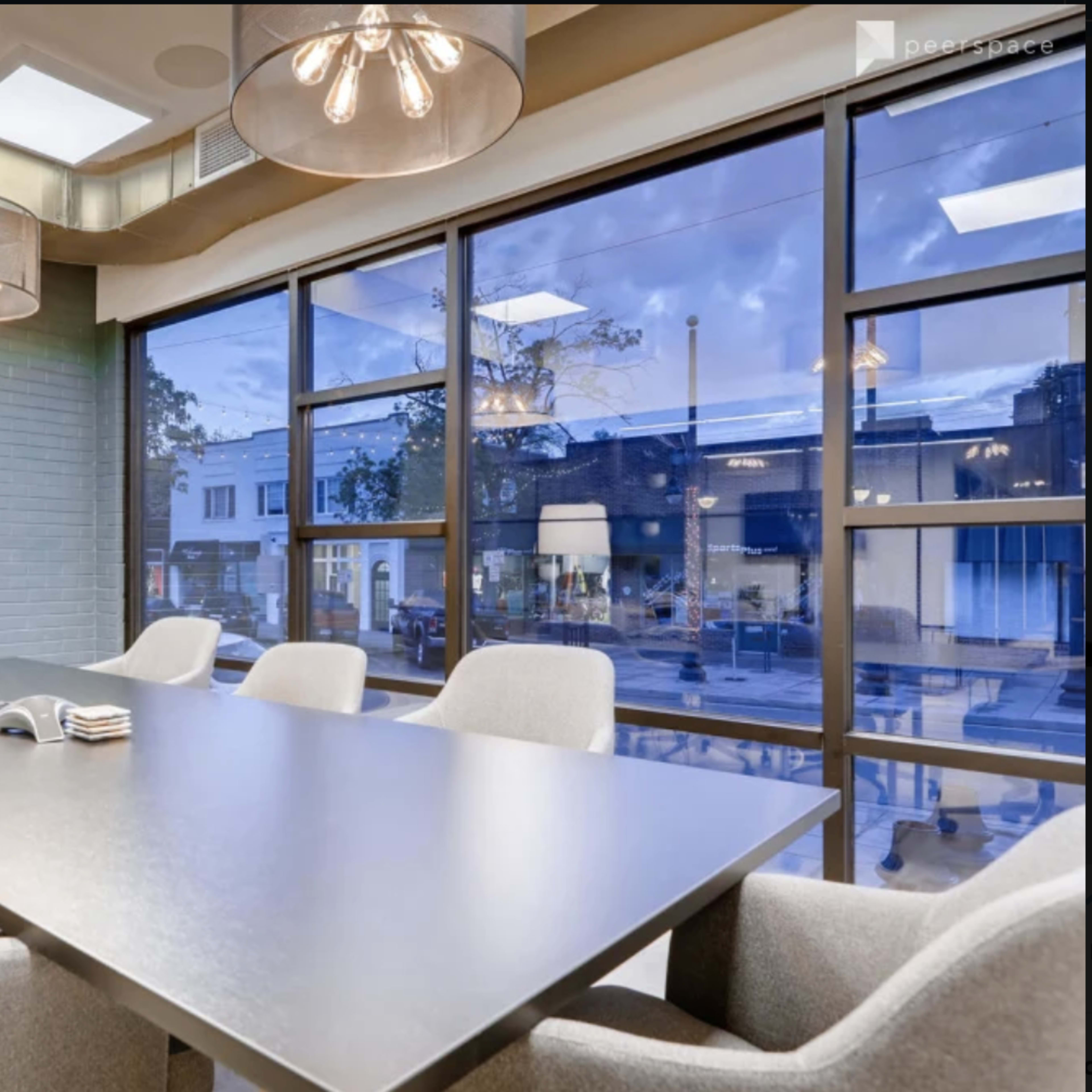 A modern conference room features a long table surrounded by gray chairs, with large windows displaying an evening cityscape outside.