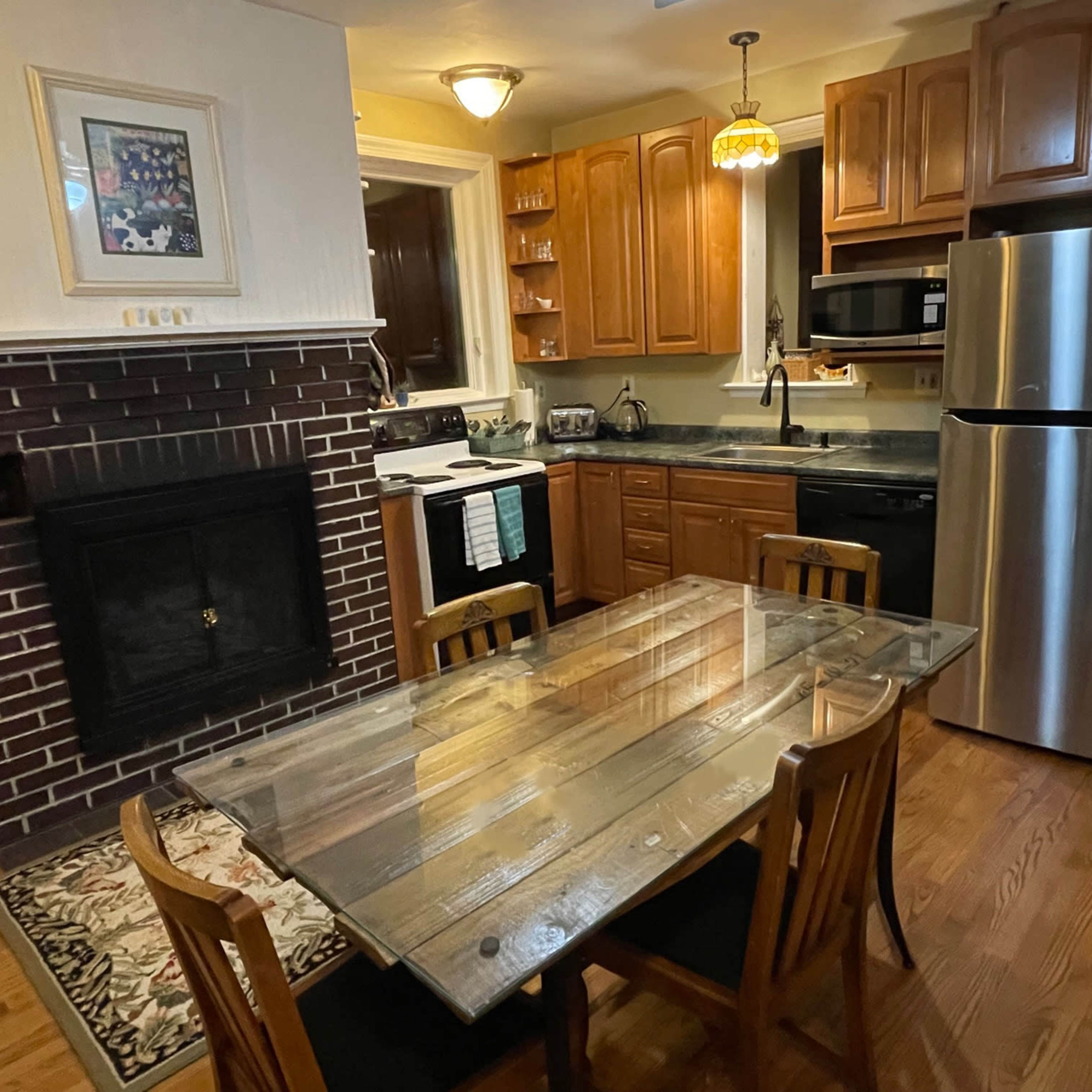 A kitchen with wooden cabinetry, a stainless steel refrigerator, a glass-top dining table with four chairs, and a fireplace on the wall.