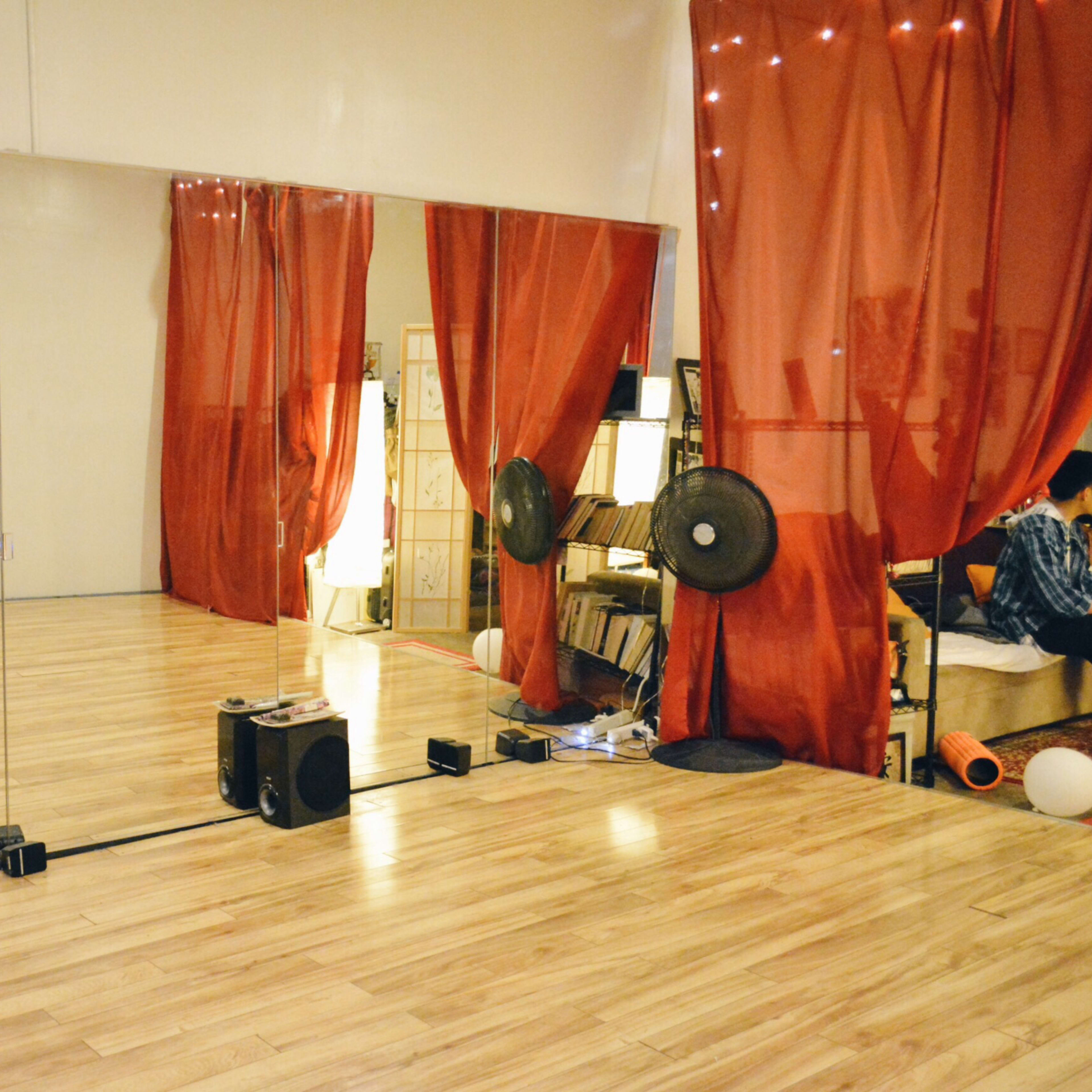 A spacious dance studio with wooden flooring, a large mirror on one wall, and red curtains, along with shelves and fans in the corner.