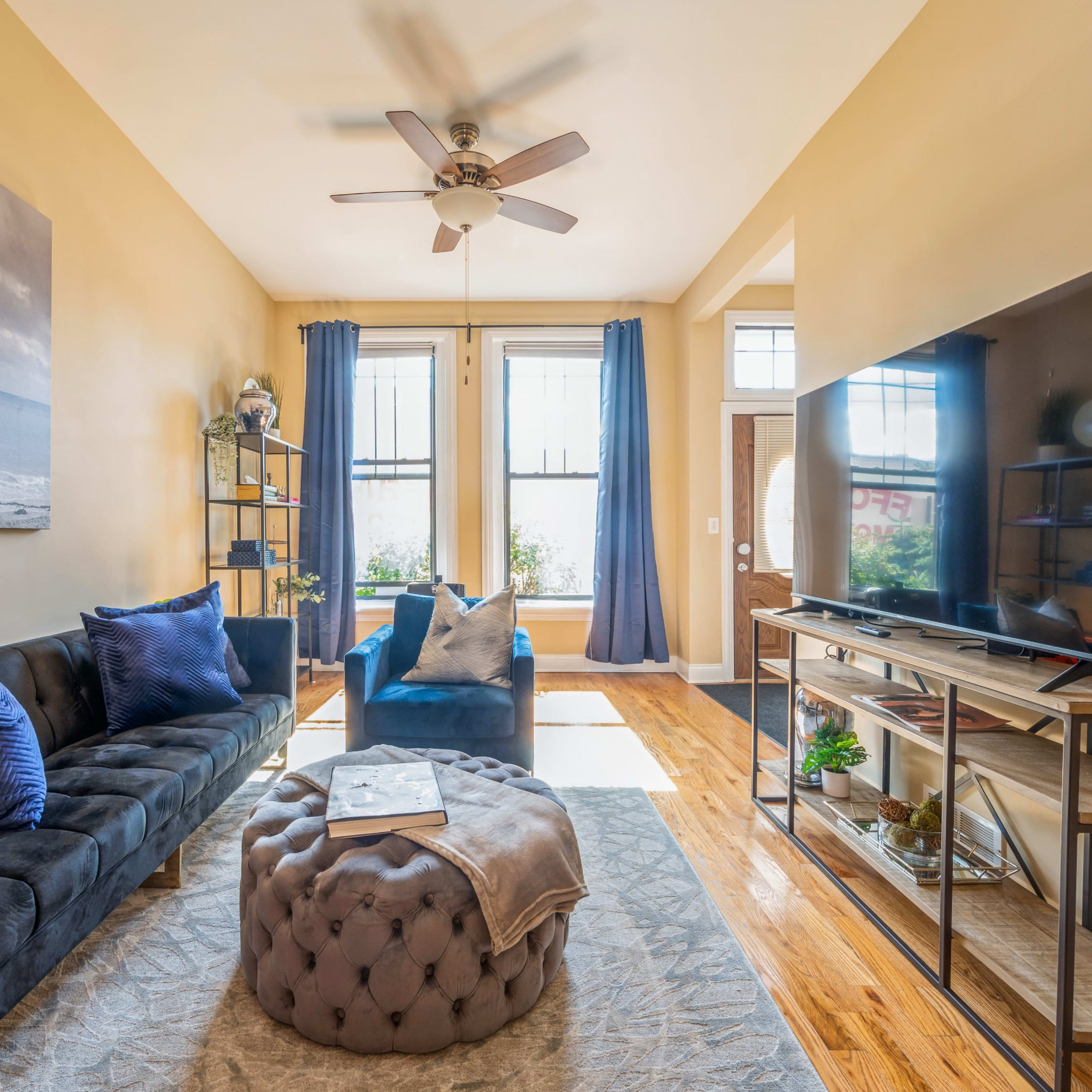 A brightly lit living room with a blue sofa, a decorative ottoman, and a large television on a stand against a wall.