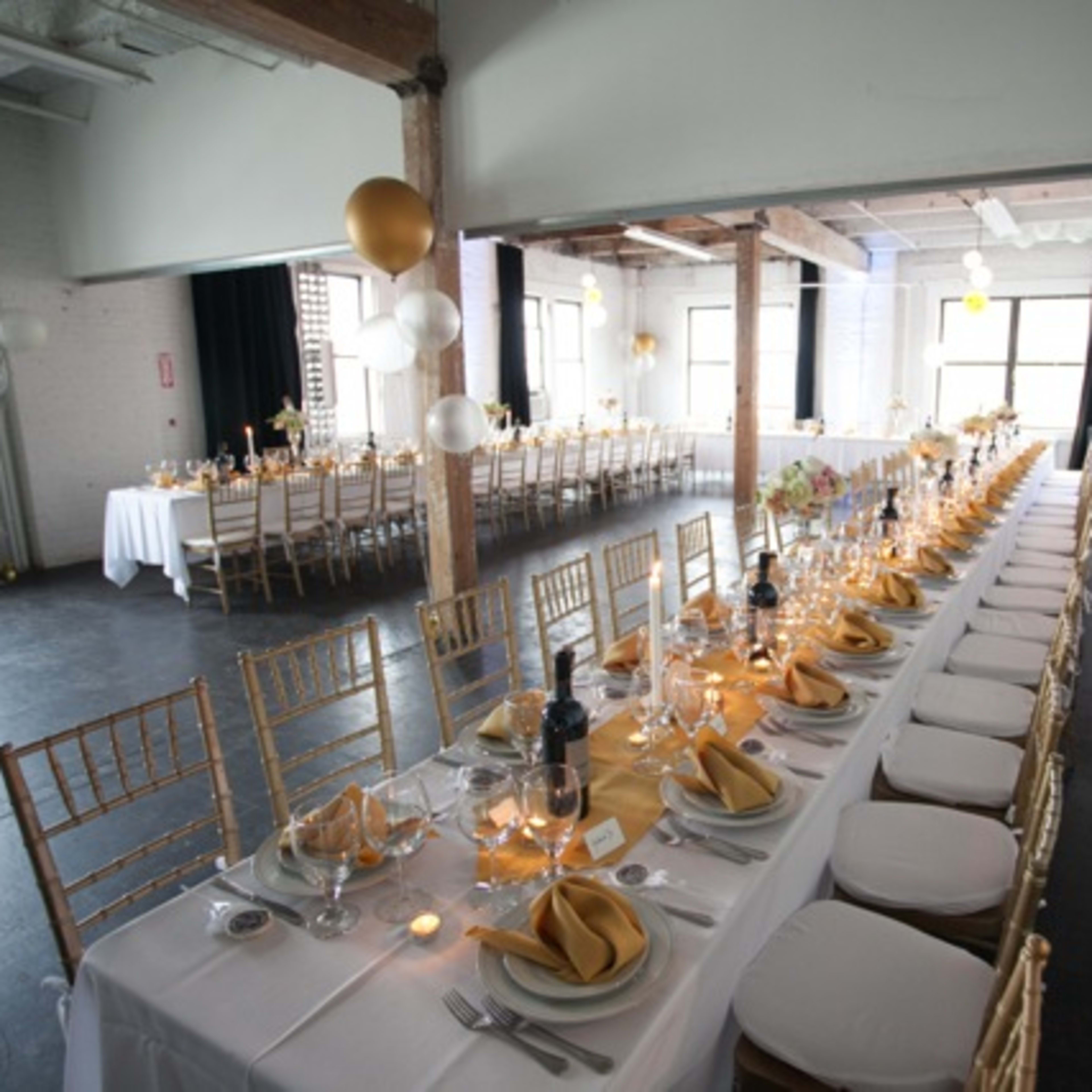 A long banquet table is set for an event inside a spacious venue, featuring gold and white decorations, with additional seating arranged in the background.
