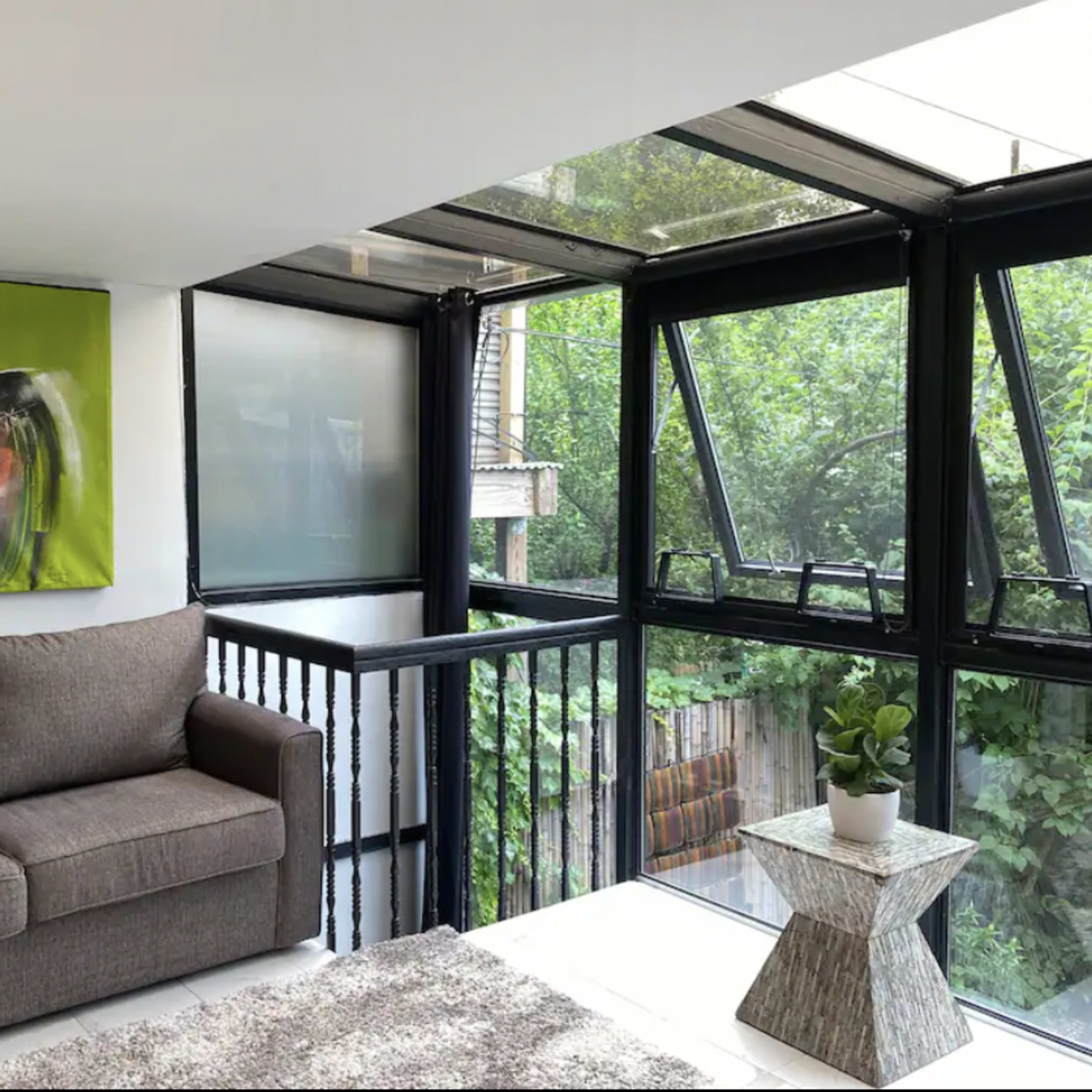 The interior features a gray sofa facing a large window, accompanied by a modern coffee table and a colorful abstract painting on the wall.