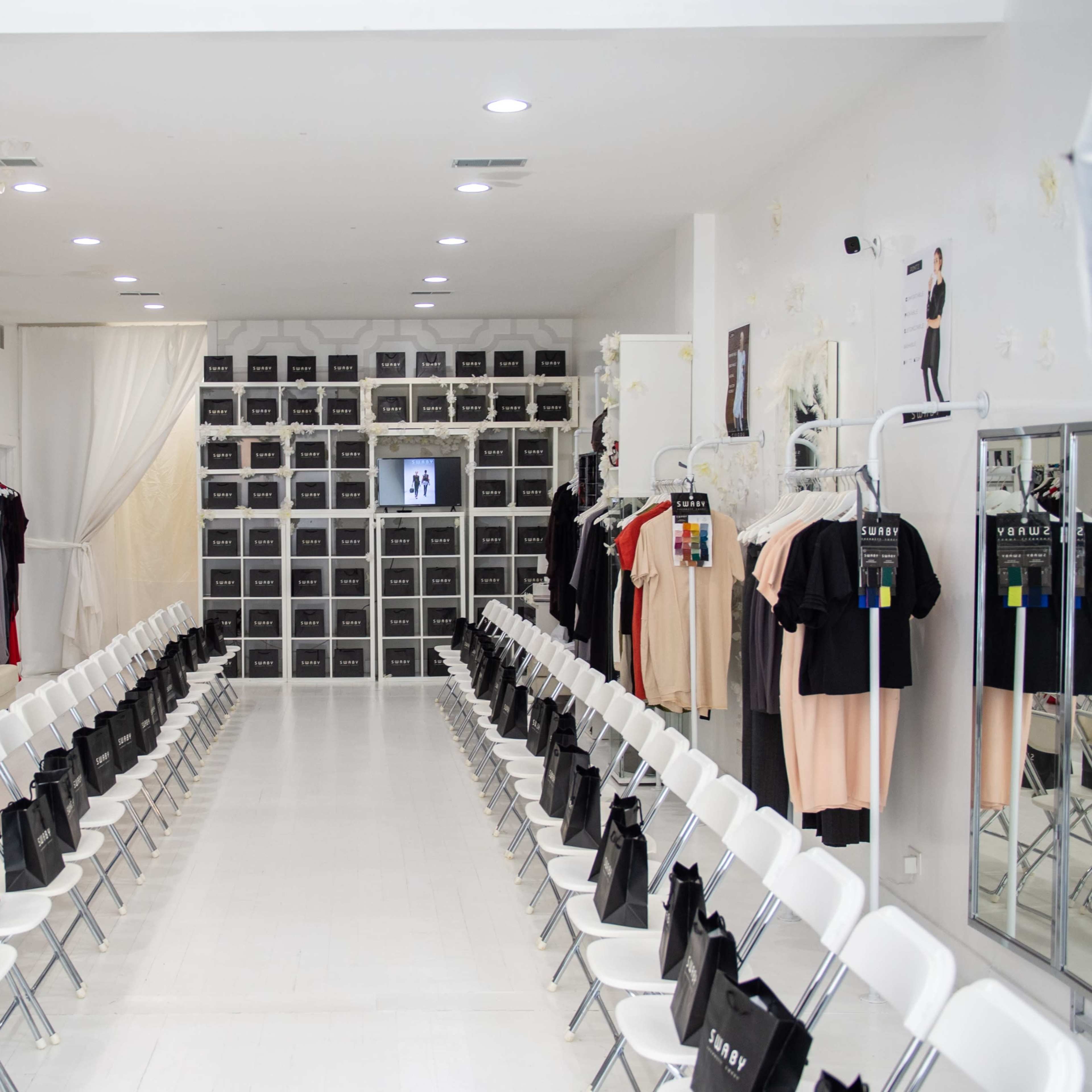 A fashion showroom with two rows of white chairs facing a presentation area flanked by clothing racks and storage units.