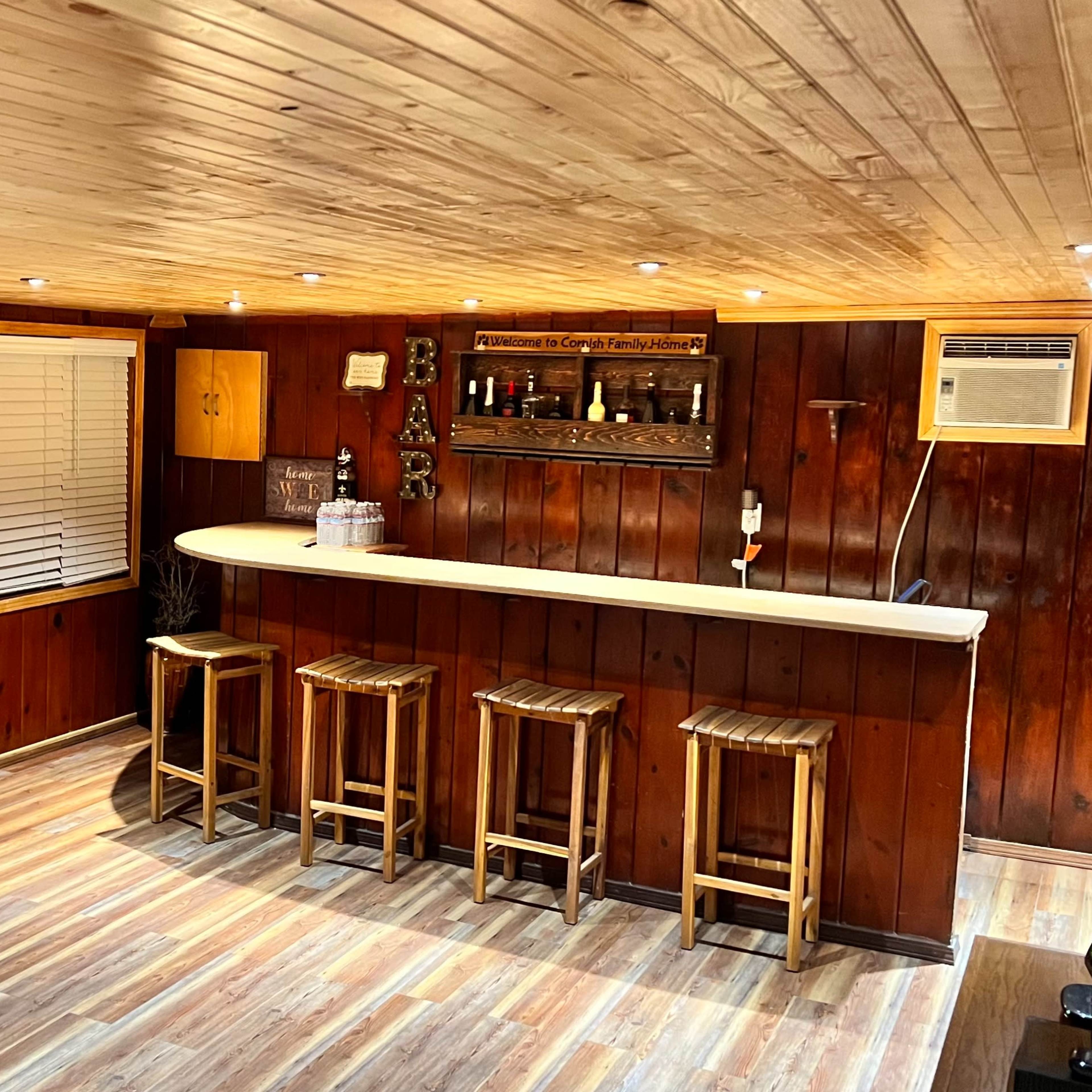 A wooden-paneled bar area with a countertop and three stools, featuring shelves with bottles in the background.