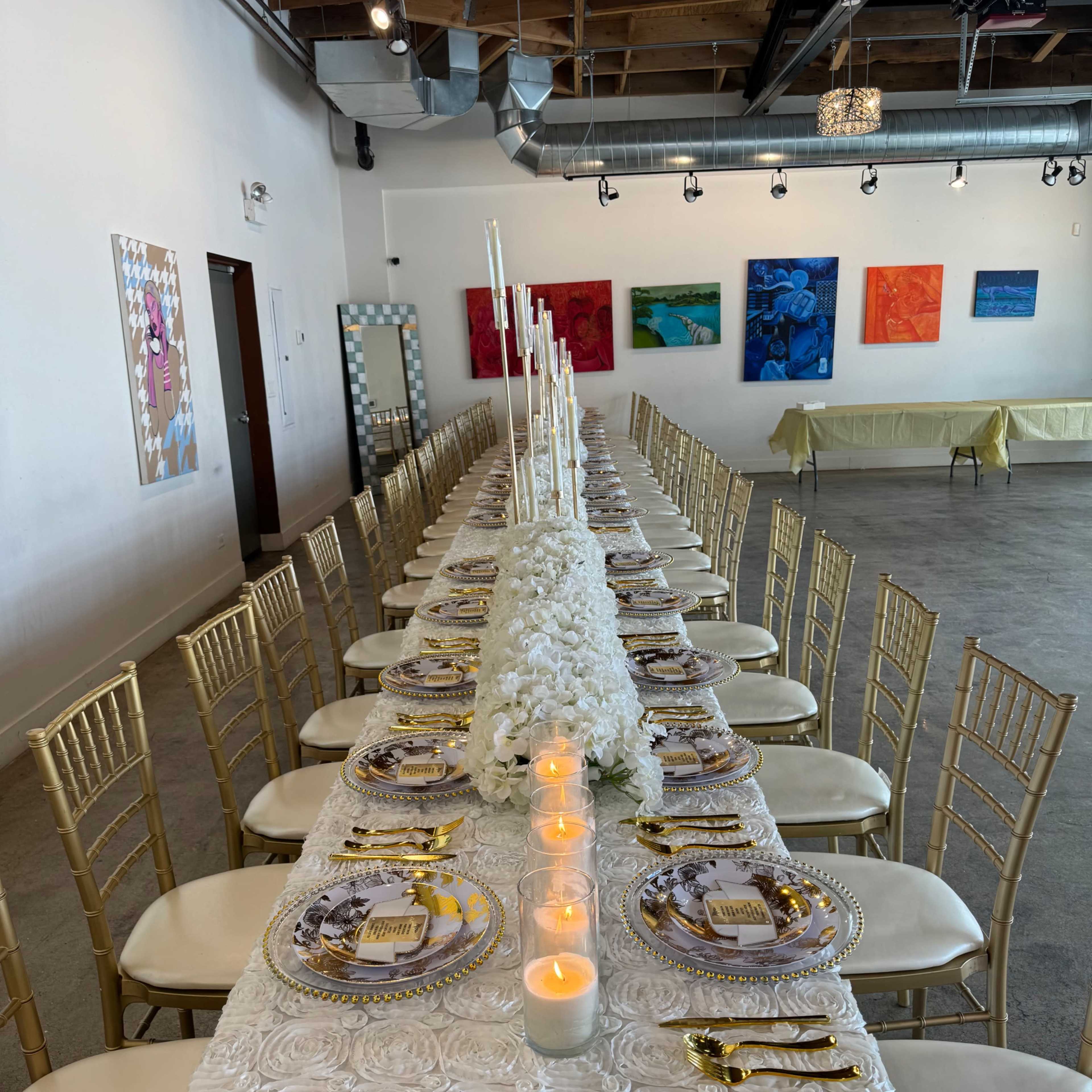 A long dining table is set with white linens, gold chairs, and plates, illuminated by candles and surrounded by colorful artworks on the walls.
