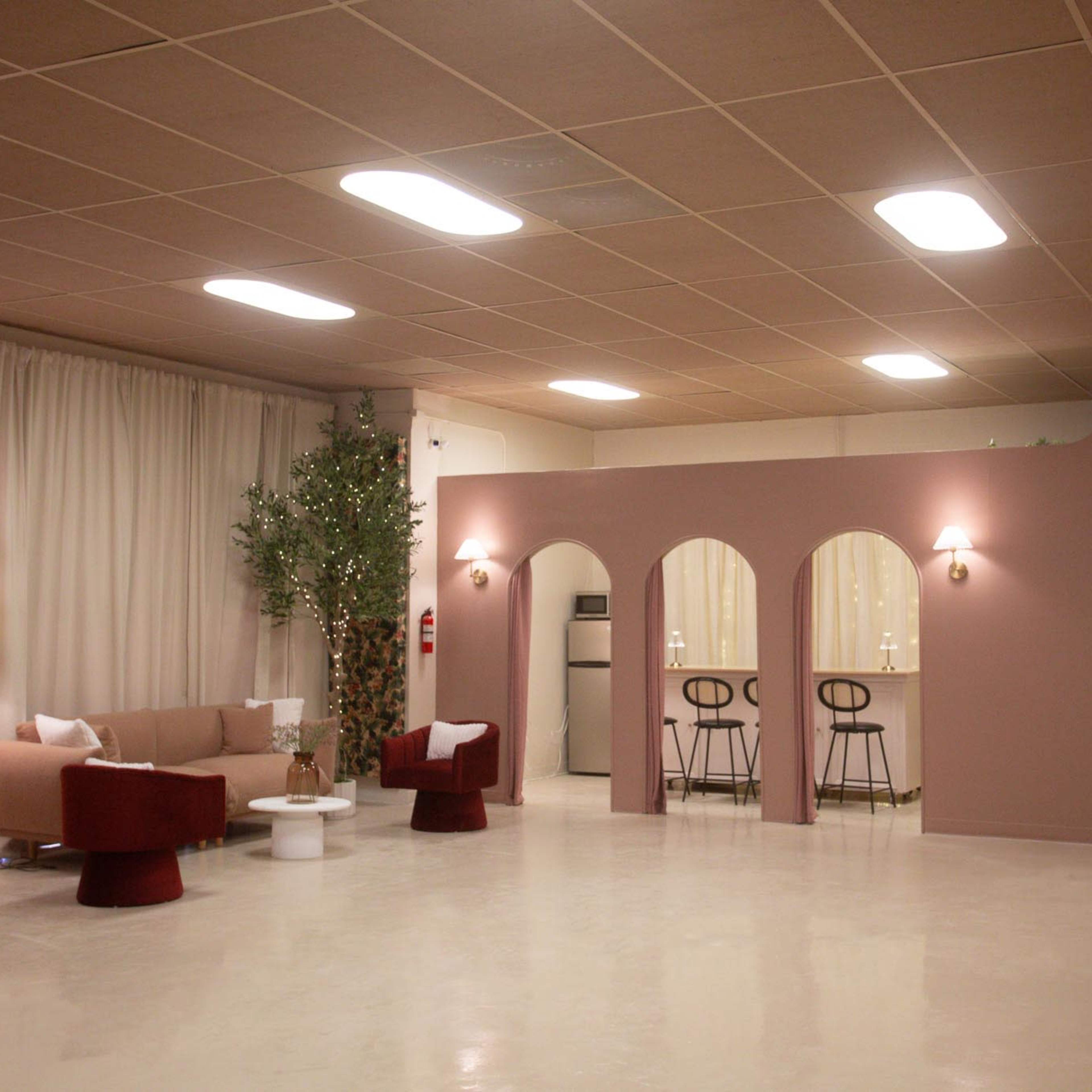 A modern indoor space with a seating area, featuring a beige sofa, red chairs, and a small table, alongside a bar area with stools against a pink wall.