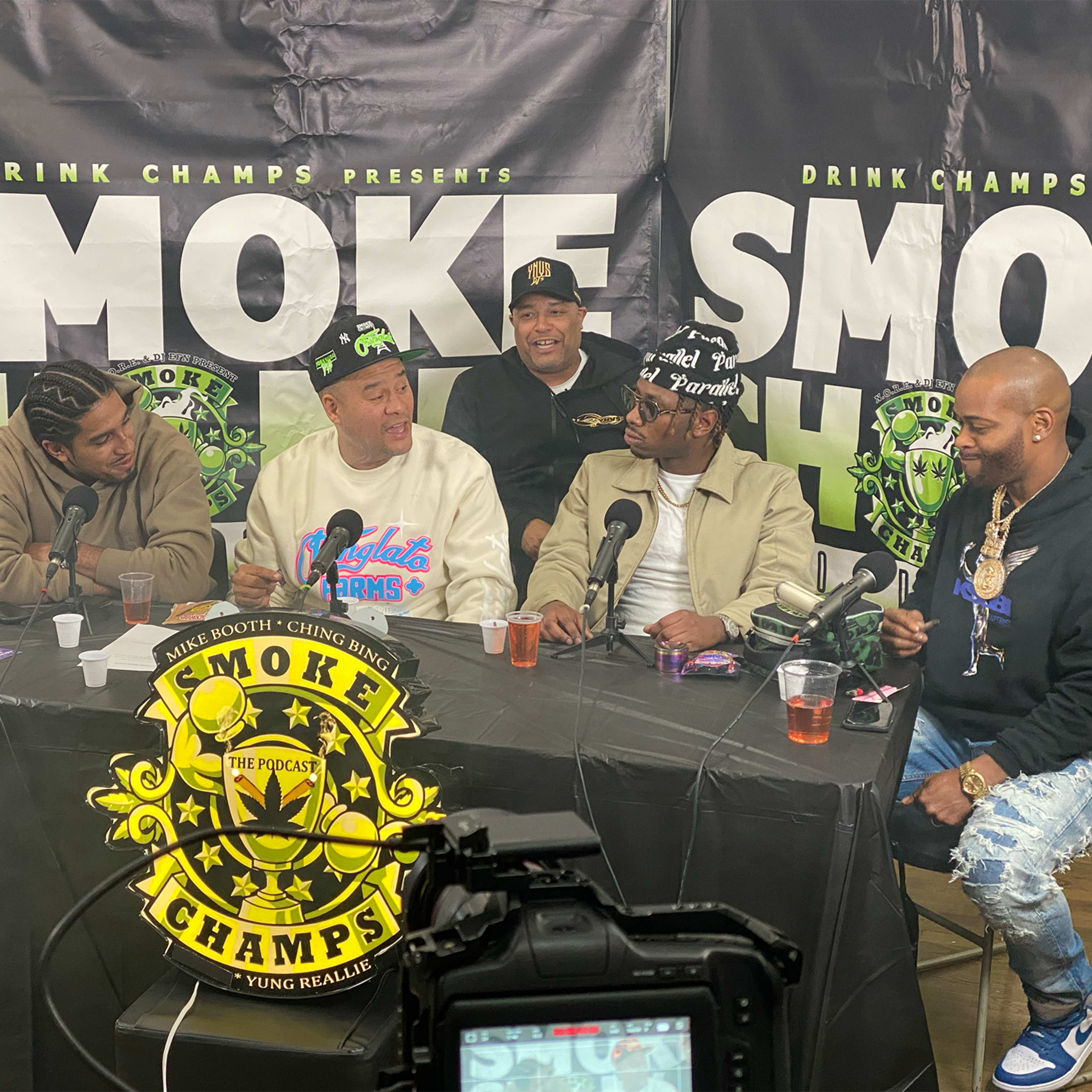 A group of five men sits around a table discussing a topic, with a branded backdrop promoting "Smoke Champs."
