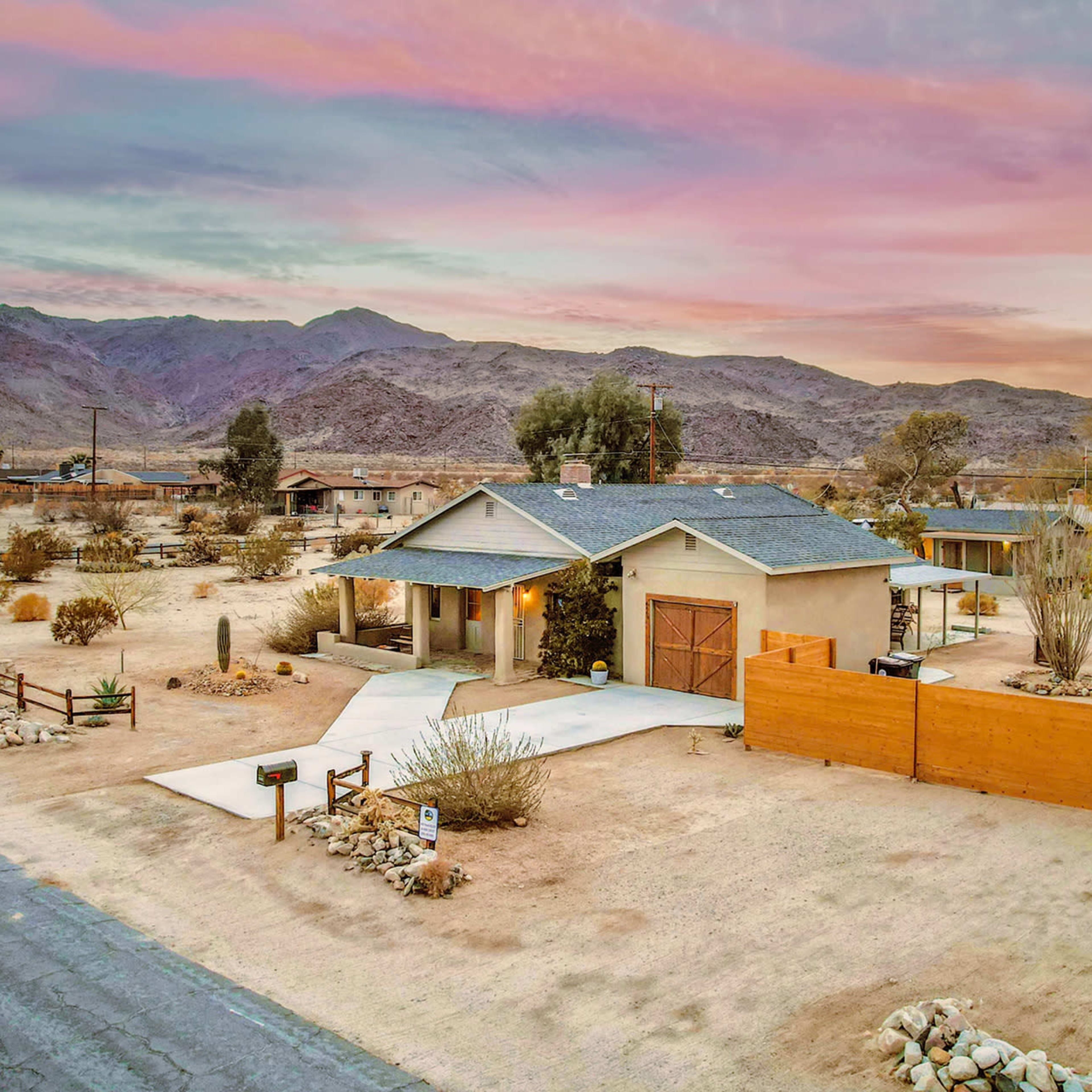 A single-story house sits on a desert landscape surrounded by mountains, with a gravel driveway and wooden fence.