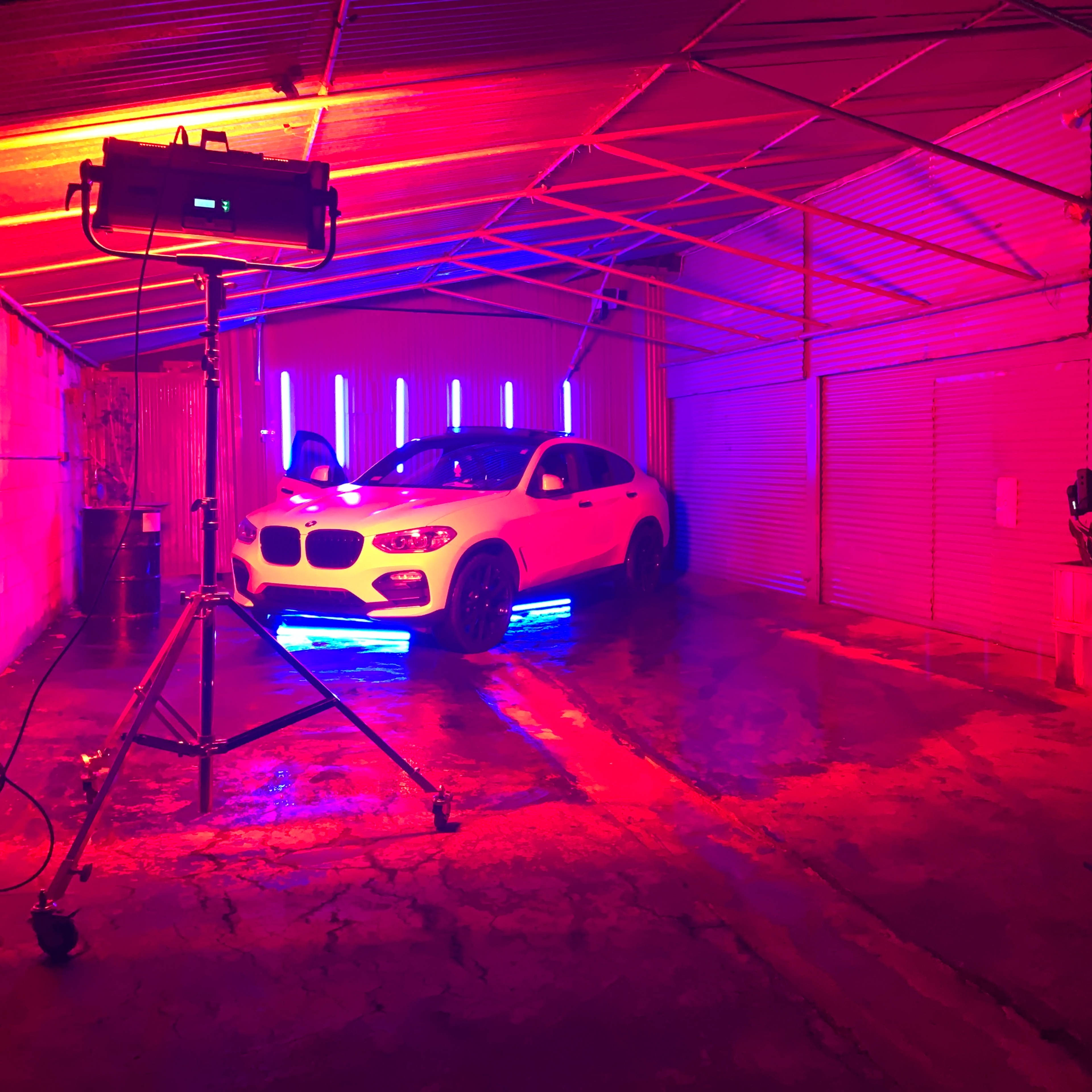 A white car is positioned in a dimly lit garage with colorful neon lights, while a person operates a camera nearby.