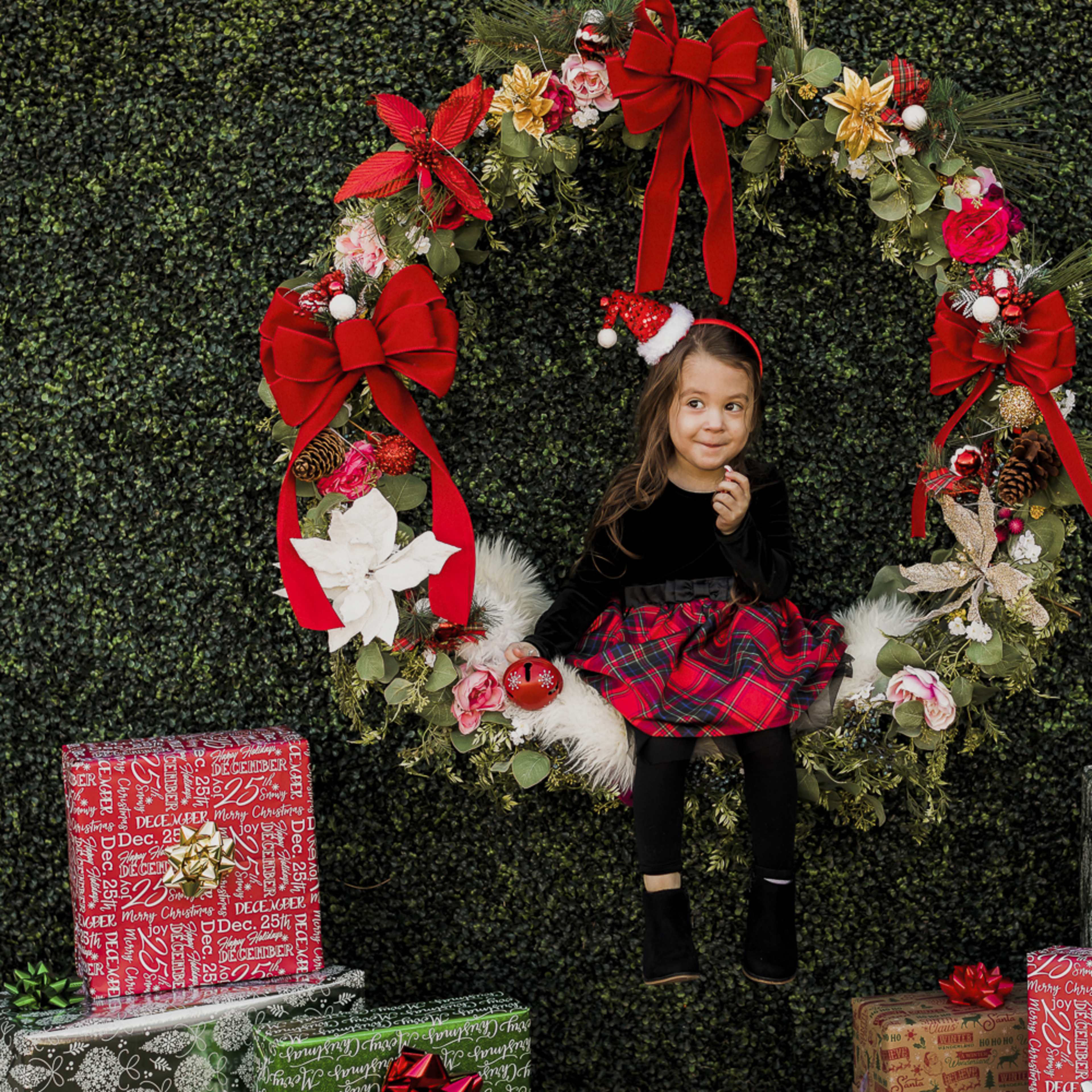 A young girl in a black top and plaid skirt sits inside a festive wreath surrounded by wrapped Christmas presents.