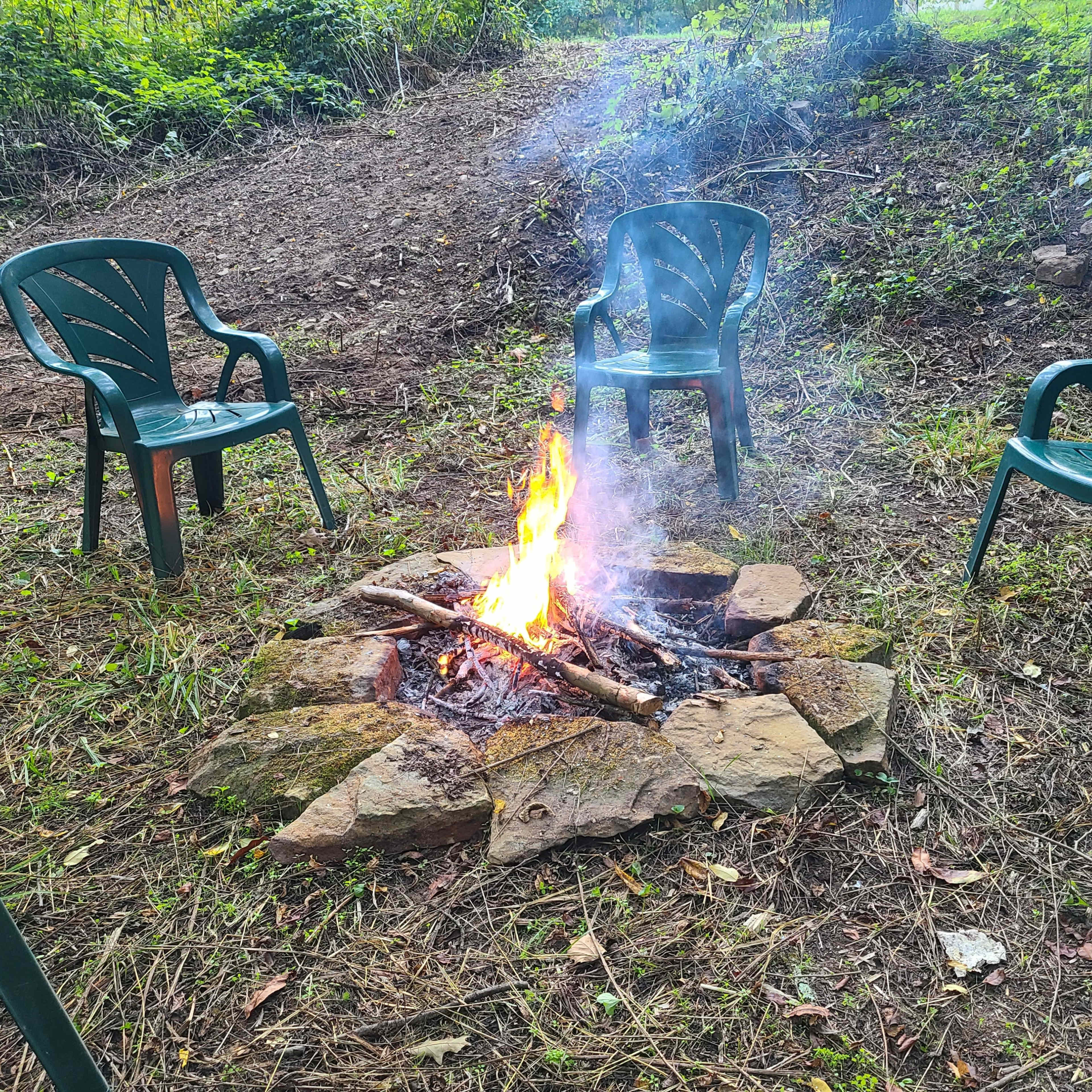 A small campfire burns in a stone circle surrounded by four green chairs in a wooded area.